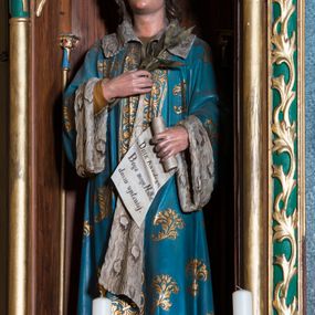 Zdjęcie nr 1: Rzeźba pełna na cokole przedstawiająca stojącego frontalnie św. Kazimierza. Święty ma lekko uniesioną głowę, oczy skierowane w górę, nos prosty, usta wąskie. Jasne włosy spływają mu falami na plecy, na głowie mitra książęca. Prawą ręką przyciska do piersi palmę męczeństwa, lewą zgiętą w łokciu trzyma rozwijający się rotulus z napisem „Dnia Każdego, / Boga mego„ Matkę, / duszo wysławiaj.” Święty jest ubrany w niebieski płaszcz zdobiony złotymi motywami roślinnymi, podbity białym futrem, spod niego wystaje jasnobrązowa suknia oraz wysokie brązowe buty. Polichromia w partiach ciała naturalistyczna.