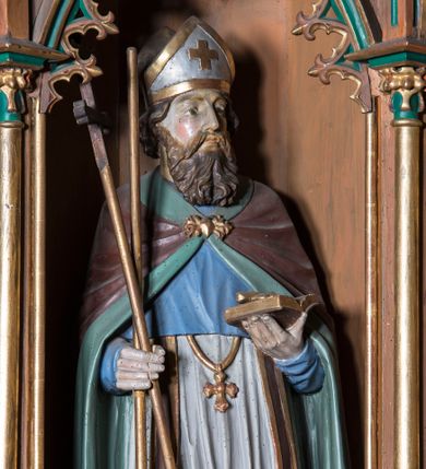 Zdjęcie nr 1: Rzeźba pełna na cokole przedstawiająca św. Wojciecha zwróconego nieznacznie w lewą stronę. Święty odwraca głowę w lewo, oczy ma skierowane przed siebie, nos prosty, usta wąskie, wydatne kości policzkowe. Głowę okala bujny brązowy zarost oraz kręcone włosy, które spływają na kark; na głowie infuła. Wojciech prawą ręką podtrzymuje wiosło oraz krzyż, w lewej trzyma otwartą księgę. Jest ubrany w ciemnobrązową albę, białą komżę ze złoconą koronką u dołu, ciemnobrązową stułę ze złoconymi brzegami, złoty krzyż biskupi i brązowy płaszcz z zielonym podszyciem, który jest spięty na piersi złotą fibulą. Spod szat widoczne ciemnobrązowe buty. Polichromia w partiach ciała naturalistyczna.
