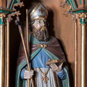 Zdjęcie nr 1: Rzeźba pełna na cokole przedstawiająca św. Wojciecha zwróconego nieznacznie w lewą stronę. Święty odwraca głowę w lewo, oczy ma skierowane przed siebie, nos prosty, usta wąskie, wydatne kości policzkowe. Głowę okala bujny brązowy zarost oraz kręcone włosy, które spływają na kark; na głowie infuła. Wojciech prawą ręką podtrzymuje wiosło oraz krzyż, w lewej trzyma otwartą księgę. Jest ubrany w ciemnobrązową albę, białą komżę ze złoconą koronką u dołu, ciemnobrązową stułę ze złoconymi brzegami, złoty krzyż biskupi i brązowy płaszcz z zielonym podszyciem, który jest spięty na piersi złotą fibulą. Spod szat widoczne ciemnobrązowe buty. Polichromia w partiach ciała naturalistyczna.
