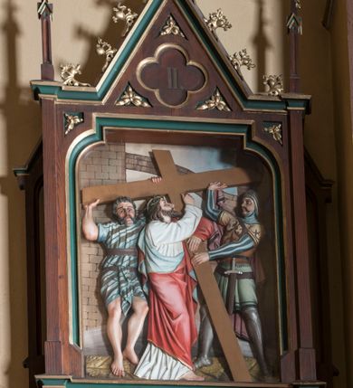 Zdjęcie nr 2: Stacje drogi krzyżowej wkomponowane w rozbudowane, drewniane neogotyckie ramy o formach przypominających kapliczki, flankowane uszakami w kształcie przypór oraz zwieńczone parami pinakli i wimpergą dekorowaną żabkami i zakończoną kwiatonem z krzyżem. Wpisano w nie motyw czworoliścia otoczony trzema rozetkami. Od dołu dekoracyjne podwieszenie. Poszczególne przedstawienia umieszczone za szybą, ujętą w profilowane obramienie w kształcie stojącego prostokąta zamkniętego łukiem nadwieszonym. Sceny są płaskorzeźbione w głębokim reliefie i ukazane na malowanym tle. Dominują zwarte, wielopostaciowe kompozycje o ekspresji wyrażonej zarówno w gestach postaci, jak i mimice twarzy. Kolorystyka pastelowa, dominują odcienie brązu, bieli i niebieskiego ożywione akcentami intensywnej czerwieni płaszcza Chrystusa. W tle fragmenty zabudowań oraz niebo, przybierające barwę od jasnego błękitu po intensywnie niebieski. W czworoliściu wimpergi numery stacji wyrażone cyframi rzymskimi, w dolnej partii obramienia prostokątne płyciny z nazwami stacji:
„I : P(an) Jezus przez Piłata na śmierć skazany”,
„II: P(an) Jezus obciążony krzyżem”, „III: P(an) Jezus upada pierwszy raz”, „IV: P(an) Jezus spotyka swą Matkę”, „V: P(an) Jezusowi dopomaga Szymon Cyre”, „VI: P(an) Jezusowi ociera twarz S. Weronika”, „VII: P(an) Jezus upada drugi raz”, „VIII: P(an) Jezus pociesza płaczące niewiasty”, „IX: P(an) Jezus upada po raz trzeci”, „X: P(an) Jezus obnażony żółcią napawany”, „XI: P(an) Jezus do krzyża przybijany”, „XII: P(an) Jezus umiera na krzyżu”, „XIII: P(an) Jezus zdjęty z krzyża”
„XIV: P(an) Jezus złożony w grobie”. 
