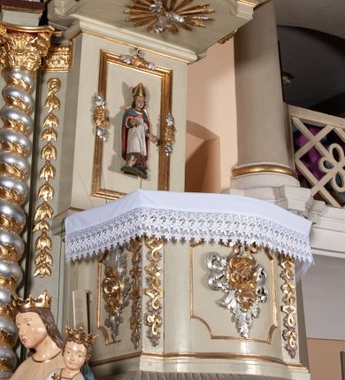 Zdjęcie nr 4: Ambona przyścienna, wisząca, z dostępem od zakrystii. Kosz na rzucie prostokąta z wypukłą ścianką frontową, z dodatkowym podestem od strony prezbiterium, ustawiony na niewielkiej podstawie dekorowanej muszlą, z podwieszeniem w formie zwężającego się cokołu ozdobionego ornamentem roślinnym, zakończonym szyszką. Ścianki kosza dekorowane płycinami w kształcie kwadratu o wklęsłych narożach wypełnionymi dekoracją złożoną z akantu, rozet i kratki regencyjnej. Naroża ścianki przedniej zdobione zwisami utworzonymi z wstęgi i kratki regencyjnej oraz ornamentu roślinnego. Parapet w formie gzymsu. Zaplecek w kształcie pionowego prostokąta, dekorowany w polu profilowaną płyciną z ornamentem roślinnym i płaskorzeźbioną figurą św. Stanisława. Baldachim powtarzający plan kosza, ujęty niepełnym belkowaniem, dekorowanym z trzech stron szczytami, z fragmentami zwróconego na zewnątrz przerwanego, wolutowego przyczółka, stanowiący podstawę dla figury anioła. Na podniebieniu baldachimu gołębica Ducha Świętego w glorii promienistej. Struktura polichromowana w kolorze jasnozielonym ze złoconymi i srebrzonymi profilami oraz ornamentyką. 
Figura anioła pełnoplastyczna, zwrócona jest w trzech czwartych w prawo, w silnym kontrapoście, o esowato wygiętej sylwetce, z głową skierowaną w górę. Obiema rękami obejmuje trąbę, na której gra. Twarz ma pociągłą, o dużych oczach skierowanych w górę, zadartym nosie i pełnych ustach, okoloną silnie kręconymi włosami. Anioł ubrany jest w zapinaną z przodu na guziki, obcisłą kamizelkę z bufiastymi, krótkimi rękawami i wysokim kołnierzem, w pludry i pończochy, wokół bioder ma zawieszony płaszcz, podwiązany po bokach. Skrzydła duże, złożone po bokach postaci. Polichromia ciała naturalistyczna, strój złocony, ze srebrzonymi detalami. 
Po wewnętrznej stronie fragmentu lewego przerwanego przyczółka napis: „odnowiono 1841. / ołtarze 3. tęcę i ambonę / przez Malarza z Tar/nawy Andrzeja / Wierzch[…]”.