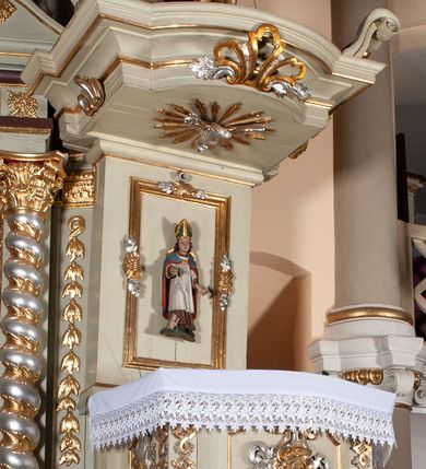 Zdjęcie nr 5: Ambona przyścienna, wisząca, z dostępem od zakrystii. Kosz na rzucie prostokąta z wypukłą ścianką frontową, z dodatkowym podestem od strony prezbiterium, ustawiony na niewielkiej podstawie dekorowanej muszlą, z podwieszeniem w formie zwężającego się cokołu ozdobionego ornamentem roślinnym, zakończonym szyszką. Ścianki kosza dekorowane płycinami w kształcie kwadratu o wklęsłych narożach wypełnionymi dekoracją złożoną z akantu, rozet i kratki regencyjnej. Naroża ścianki przedniej zdobione zwisami utworzonymi z wstęgi i kratki regencyjnej oraz ornamentu roślinnego. Parapet w formie gzymsu. Zaplecek w kształcie pionowego prostokąta, dekorowany w polu profilowaną płyciną z ornamentem roślinnym i płaskorzeźbioną figurą św. Stanisława. Baldachim powtarzający plan kosza, ujęty niepełnym belkowaniem, dekorowanym z trzech stron szczytami, z fragmentami zwróconego na zewnątrz przerwanego, wolutowego przyczółka, stanowiący podstawę dla figury anioła. Na podniebieniu baldachimu gołębica Ducha Świętego w glorii promienistej. Struktura polichromowana w kolorze jasnozielonym ze złoconymi i srebrzonymi profilami oraz ornamentyką. 
Figura anioła pełnoplastyczna, zwrócona jest w trzech czwartych w prawo, w silnym kontrapoście, o esowato wygiętej sylwetce, z głową skierowaną w górę. Obiema rękami obejmuje trąbę, na której gra. Twarz ma pociągłą, o dużych oczach skierowanych w górę, zadartym nosie i pełnych ustach, okoloną silnie kręconymi włosami. Anioł ubrany jest w zapinaną z przodu na guziki, obcisłą kamizelkę z bufiastymi, krótkimi rękawami i wysokim kołnierzem, w pludry i pończochy, wokół bioder ma zawieszony płaszcz, podwiązany po bokach. Skrzydła duże, złożone po bokach postaci. Polichromia ciała naturalistyczna, strój złocony, ze srebrzonymi detalami. 
Po wewnętrznej stronie fragmentu lewego przerwanego przyczółka napis: „odnowiono 1841. / ołtarze 3. tęcę i ambonę / przez Malarza z Tar/nawy Andrzeja / Wierzch[…]”.