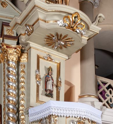 Zdjęcie nr 1: Ambona przyścienna, wisząca, z dostępem od zakrystii. Kosz na rzucie prostokąta z wypukłą ścianką frontową, z dodatkowym podestem od strony prezbiterium, ustawiony na niewielkiej podstawie dekorowanej muszlą, z podwieszeniem w formie zwężającego się cokołu ozdobionego ornamentem roślinnym, zakończonym szyszką. Ścianki kosza dekorowane płycinami w kształcie kwadratu o wklęsłych narożach wypełnionymi dekoracją złożoną z akantu, rozet i kratki regencyjnej. Naroża ścianki przedniej zdobione zwisami utworzonymi z wstęgi i kratki regencyjnej oraz ornamentu roślinnego. Parapet w formie gzymsu. Zaplecek w kształcie pionowego prostokąta, dekorowany w polu profilowaną płyciną z ornamentem roślinnym i płaskorzeźbioną figurą św. Stanisława. Baldachim powtarzający plan kosza, ujęty niepełnym belkowaniem, dekorowanym z trzech stron szczytami, z fragmentami zwróconego na zewnątrz przerwanego, wolutowego przyczółka, stanowiący podstawę dla figury anioła. Na podniebieniu baldachimu gołębica Ducha Świętego w glorii promienistej. Struktura polichromowana w kolorze jasnozielonym ze złoconymi i srebrzonymi profilami oraz ornamentyką. 
Figura anioła pełnoplastyczna, zwrócona jest w trzech czwartych w prawo, w silnym kontrapoście, o esowato wygiętej sylwetce, z głową skierowaną w górę. Obiema rękami obejmuje trąbę, na której gra. Twarz ma pociągłą, o dużych oczach skierowanych w górę, zadartym nosie i pełnych ustach, okoloną silnie kręconymi włosami. Anioł ubrany jest w zapinaną z przodu na guziki, obcisłą kamizelkę z bufiastymi, krótkimi rękawami i wysokim kołnierzem, w pludry i pończochy, wokół bioder ma zawieszony płaszcz, podwiązany po bokach. Skrzydła duże, złożone po bokach postaci. Polichromia ciała naturalistyczna, strój złocony, ze srebrzonymi detalami. 
Po wewnętrznej stronie fragmentu lewego przerwanego przyczółka napis: „odnowiono 1841. / ołtarze 3. tęcę i ambonę / przez Malarza z Tar/nawy Andrzeja / Wierzch[…]”.