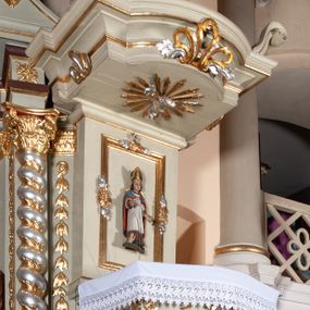 Zdjęcie nr 1: Ambona przyścienna, wisząca, z dostępem od zakrystii. Kosz na rzucie prostokąta z wypukłą ścianką frontową, z dodatkowym podestem od strony prezbiterium, ustawiony na niewielkiej podstawie dekorowanej muszlą, z podwieszeniem w formie zwężającego się cokołu ozdobionego ornamentem roślinnym, zakończonym szyszką. Ścianki kosza dekorowane płycinami w kształcie kwadratu o wklęsłych narożach wypełnionymi dekoracją złożoną z akantu, rozet i kratki regencyjnej. Naroża ścianki przedniej zdobione zwisami utworzonymi z wstęgi i kratki regencyjnej oraz ornamentu roślinnego. Parapet w formie gzymsu. Zaplecek w kształcie pionowego prostokąta, dekorowany w polu profilowaną płyciną z ornamentem roślinnym i płaskorzeźbioną figurą św. Stanisława. Baldachim powtarzający plan kosza, ujęty niepełnym belkowaniem, dekorowanym z trzech stron szczytami, z fragmentami zwróconego na zewnątrz przerwanego, wolutowego przyczółka, stanowiący podstawę dla figury anioła. Na podniebieniu baldachimu gołębica Ducha Świętego w glorii promienistej. Struktura polichromowana w kolorze jasnozielonym ze złoconymi i srebrzonymi profilami oraz ornamentyką. 
Figura anioła pełnoplastyczna, zwrócona jest w trzech czwartych w prawo, w silnym kontrapoście, o esowato wygiętej sylwetce, z głową skierowaną w górę. Obiema rękami obejmuje trąbę, na której gra. Twarz ma pociągłą, o dużych oczach skierowanych w górę, zadartym nosie i pełnych ustach, okoloną silnie kręconymi włosami. Anioł ubrany jest w zapinaną z przodu na guziki, obcisłą kamizelkę z bufiastymi, krótkimi rękawami i wysokim kołnierzem, w pludry i pończochy, wokół bioder ma zawieszony płaszcz, podwiązany po bokach. Skrzydła duże, złożone po bokach postaci. Polichromia ciała naturalistyczna, strój złocony, ze srebrzonymi detalami. 
Po wewnętrznej stronie fragmentu lewego przerwanego przyczółka napis: „odnowiono 1841. / ołtarze 3. tęcę i ambonę / przez Malarza z Tar/nawy Andrzeja / Wierzch[…]”.