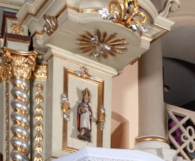 Zdjęcie nr 1: Ambona przyścienna, wisząca, z dostępem od zakrystii. Kosz na rzucie prostokąta z wypukłą ścianką frontową, z dodatkowym podestem od strony prezbiterium, ustawiony na niewielkiej podstawie dekorowanej muszlą, z podwieszeniem w formie zwężającego się cokołu ozdobionego ornamentem roślinnym, zakończonym szyszką. Ścianki kosza dekorowane płycinami w kształcie kwadratu o wklęsłych narożach wypełnionymi dekoracją złożoną z akantu, rozet i kratki regencyjnej. Naroża ścianki przedniej zdobione zwisami utworzonymi z wstęgi i kratki regencyjnej oraz ornamentu roślinnego. Parapet w formie gzymsu. Zaplecek w kształcie pionowego prostokąta, dekorowany w polu profilowaną płyciną z ornamentem roślinnym i płaskorzeźbioną figurą św. Stanisława. Baldachim powtarzający plan kosza, ujęty niepełnym belkowaniem, dekorowanym z trzech stron szczytami, z fragmentami zwróconego na zewnątrz przerwanego, wolutowego przyczółka, stanowiący podstawę dla figury anioła. Na podniebieniu baldachimu gołębica Ducha Świętego w glorii promienistej. Struktura polichromowana w kolorze jasnozielonym ze złoconymi i srebrzonymi profilami oraz ornamentyką. 
Figura anioła pełnoplastyczna, zwrócona jest w trzech czwartych w prawo, w silnym kontrapoście, o esowato wygiętej sylwetce, z głową skierowaną w górę. Obiema rękami obejmuje trąbę, na której gra. Twarz ma pociągłą, o dużych oczach skierowanych w górę, zadartym nosie i pełnych ustach, okoloną silnie kręconymi włosami. Anioł ubrany jest w zapinaną z przodu na guziki, obcisłą kamizelkę z bufiastymi, krótkimi rękawami i wysokim kołnierzem, w pludry i pończochy, wokół bioder ma zawieszony płaszcz, podwiązany po bokach. Skrzydła duże, złożone po bokach postaci. Polichromia ciała naturalistyczna, strój złocony, ze srebrzonymi detalami. 
Po wewnętrznej stronie fragmentu lewego przerwanego przyczółka napis: „odnowiono 1841. / ołtarze 3. tęcę i ambonę / przez Malarza z Tar/nawy Andrzeja / Wierzch[…]”.