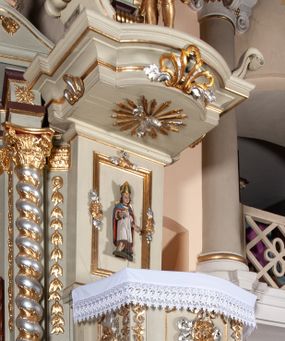 Zdjęcie nr 1: Ambona przyścienna, wisząca, z dostępem od zakrystii. Kosz na rzucie prostokąta z wypukłą ścianką frontową, z dodatkowym podestem od strony prezbiterium, ustawiony na niewielkiej podstawie dekorowanej muszlą, z podwieszeniem w formie zwężającego się cokołu ozdobionego ornamentem roślinnym, zakończonym szyszką. Ścianki kosza dekorowane płycinami w kształcie kwadratu o wklęsłych narożach wypełnionymi dekoracją złożoną z akantu, rozet i kratki regencyjnej. Naroża ścianki przedniej zdobione zwisami utworzonymi z wstęgi i kratki regencyjnej oraz ornamentu roślinnego. Parapet w formie gzymsu. Zaplecek w kształcie pionowego prostokąta, dekorowany w polu profilowaną płyciną z ornamentem roślinnym i płaskorzeźbioną figurą św. Stanisława. Baldachim powtarzający plan kosza, ujęty niepełnym belkowaniem, dekorowanym z trzech stron szczytami, z fragmentami zwróconego na zewnątrz przerwanego, wolutowego przyczółka, stanowiący podstawę dla figury anioła. Na podniebieniu baldachimu gołębica Ducha Świętego w glorii promienistej. Struktura polichromowana w kolorze jasnozielonym ze złoconymi i srebrzonymi profilami oraz ornamentyką. 
Figura anioła pełnoplastyczna, zwrócona jest w trzech czwartych w prawo, w silnym kontrapoście, o esowato wygiętej sylwetce, z głową skierowaną w górę. Obiema rękami obejmuje trąbę, na której gra. Twarz ma pociągłą, o dużych oczach skierowanych w górę, zadartym nosie i pełnych ustach, okoloną silnie kręconymi włosami. Anioł ubrany jest w zapinaną z przodu na guziki, obcisłą kamizelkę z bufiastymi, krótkimi rękawami i wysokim kołnierzem, w pludry i pończochy, wokół bioder ma zawieszony płaszcz, podwiązany po bokach. Skrzydła duże, złożone po bokach postaci. Polichromia ciała naturalistyczna, strój złocony, ze srebrzonymi detalami. 
Po wewnętrznej stronie fragmentu lewego przerwanego przyczółka napis: „odnowiono 1841. / ołtarze 3. tęcę i ambonę / przez Malarza z Tar/nawy Andrzeja / Wierzch[…]”.