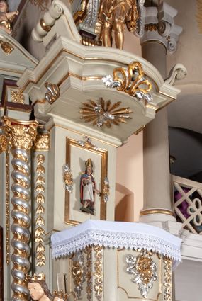 Zdjęcie nr 1: Ambona przyścienna, wisząca, z dostępem od zakrystii. Kosz na rzucie prostokąta z wypukłą ścianką frontową, z dodatkowym podestem od strony prezbiterium, ustawiony na niewielkiej podstawie dekorowanej muszlą, z podwieszeniem w formie zwężającego się cokołu ozdobionego ornamentem roślinnym, zakończonym szyszką. Ścianki kosza dekorowane płycinami w kształcie kwadratu o wklęsłych narożach wypełnionymi dekoracją złożoną z akantu, rozet i kratki regencyjnej. Naroża ścianki przedniej zdobione zwisami utworzonymi z wstęgi i kratki regencyjnej oraz ornamentu roślinnego. Parapet w formie gzymsu. Zaplecek w kształcie pionowego prostokąta, dekorowany w polu profilowaną płyciną z ornamentem roślinnym i płaskorzeźbioną figurą św. Stanisława. Baldachim powtarzający plan kosza, ujęty niepełnym belkowaniem, dekorowanym z trzech stron szczytami, z fragmentami zwróconego na zewnątrz przerwanego, wolutowego przyczółka, stanowiący podstawę dla figury anioła. Na podniebieniu baldachimu gołębica Ducha Świętego w glorii promienistej. Struktura polichromowana w kolorze jasnozielonym ze złoconymi i srebrzonymi profilami oraz ornamentyką. 
Figura anioła pełnoplastyczna, zwrócona jest w trzech czwartych w prawo, w silnym kontrapoście, o esowato wygiętej sylwetce, z głową skierowaną w górę. Obiema rękami obejmuje trąbę, na której gra. Twarz ma pociągłą, o dużych oczach skierowanych w górę, zadartym nosie i pełnych ustach, okoloną silnie kręconymi włosami. Anioł ubrany jest w zapinaną z przodu na guziki, obcisłą kamizelkę z bufiastymi, krótkimi rękawami i wysokim kołnierzem, w pludry i pończochy, wokół bioder ma zawieszony płaszcz, podwiązany po bokach. Skrzydła duże, złożone po bokach postaci. Polichromia ciała naturalistyczna, strój złocony, ze srebrzonymi detalami. 
Po wewnętrznej stronie fragmentu lewego przerwanego przyczółka napis: „odnowiono 1841. / ołtarze 3. tęcę i ambonę / przez Malarza z Tar/nawy Andrzeja / Wierzch[…]”.
