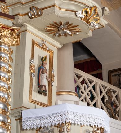 Zdjęcie nr 8: Ambona przyścienna, wisząca, z dostępem od zakrystii. Kosz na rzucie prostokąta z wypukłą ścianką frontową, z dodatkowym podestem od strony prezbiterium, ustawiony na niewielkiej podstawie dekorowanej muszlą, z podwieszeniem w formie zwężającego się cokołu ozdobionego ornamentem roślinnym, zakończonym szyszką. Ścianki kosza dekorowane płycinami w kształcie kwadratu o wklęsłych narożach wypełnionymi dekoracją złożoną z akantu, rozet i kratki regencyjnej. Naroża ścianki przedniej zdobione zwisami utworzonymi z wstęgi i kratki regencyjnej oraz ornamentu roślinnego. Parapet w formie gzymsu. Zaplecek w kształcie pionowego prostokąta, dekorowany w polu profilowaną płyciną z ornamentem roślinnym i płaskorzeźbioną figurą św. Stanisława. Baldachim powtarzający plan kosza, ujęty niepełnym belkowaniem, dekorowanym z trzech stron szczytami, z fragmentami zwróconego na zewnątrz przerwanego, wolutowego przyczółka, stanowiący podstawę dla figury anioła. Na podniebieniu baldachimu gołębica Ducha Świętego w glorii promienistej. Struktura polichromowana w kolorze jasnozielonym ze złoconymi i srebrzonymi profilami oraz ornamentyką. 
Figura anioła pełnoplastyczna, zwrócona jest w trzech czwartych w prawo, w silnym kontrapoście, o esowato wygiętej sylwetce, z głową skierowaną w górę. Obiema rękami obejmuje trąbę, na której gra. Twarz ma pociągłą, o dużych oczach skierowanych w górę, zadartym nosie i pełnych ustach, okoloną silnie kręconymi włosami. Anioł ubrany jest w zapinaną z przodu na guziki, obcisłą kamizelkę z bufiastymi, krótkimi rękawami i wysokim kołnierzem, w pludry i pończochy, wokół bioder ma zawieszony płaszcz, podwiązany po bokach. Skrzydła duże, złożone po bokach postaci. Polichromia ciała naturalistyczna, strój złocony, ze srebrzonymi detalami. 
Po wewnętrznej stronie fragmentu lewego przerwanego przyczółka napis: „odnowiono 1841. / ołtarze 3. tęcę i ambonę / przez Malarza z Tar/nawy Andrzeja / Wierzch[…]”.