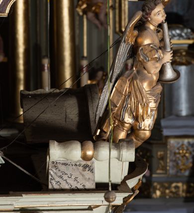 Zdjęcie nr 12: Ambona przyścienna, wisząca, z dostępem od zakrystii. Kosz na rzucie prostokąta z wypukłą ścianką frontową, z dodatkowym podestem od strony prezbiterium, ustawiony na niewielkiej podstawie dekorowanej muszlą, z podwieszeniem w formie zwężającego się cokołu ozdobionego ornamentem roślinnym, zakończonym szyszką. Ścianki kosza dekorowane płycinami w kształcie kwadratu o wklęsłych narożach wypełnionymi dekoracją złożoną z akantu, rozet i kratki regencyjnej. Naroża ścianki przedniej zdobione zwisami utworzonymi z wstęgi i kratki regencyjnej oraz ornamentu roślinnego. Parapet w formie gzymsu. Zaplecek w kształcie pionowego prostokąta, dekorowany w polu profilowaną płyciną z ornamentem roślinnym i płaskorzeźbioną figurą św. Stanisława. Baldachim powtarzający plan kosza, ujęty niepełnym belkowaniem, dekorowanym z trzech stron szczytami, z fragmentami zwróconego na zewnątrz przerwanego, wolutowego przyczółka, stanowiący podstawę dla figury anioła. Na podniebieniu baldachimu gołębica Ducha Świętego w glorii promienistej. Struktura polichromowana w kolorze jasnozielonym ze złoconymi i srebrzonymi profilami oraz ornamentyką. 
Figura anioła pełnoplastyczna, zwrócona jest w trzech czwartych w prawo, w silnym kontrapoście, o esowato wygiętej sylwetce, z głową skierowaną w górę. Obiema rękami obejmuje trąbę, na której gra. Twarz ma pociągłą, o dużych oczach skierowanych w górę, zadartym nosie i pełnych ustach, okoloną silnie kręconymi włosami. Anioł ubrany jest w zapinaną z przodu na guziki, obcisłą kamizelkę z bufiastymi, krótkimi rękawami i wysokim kołnierzem, w pludry i pończochy, wokół bioder ma zawieszony płaszcz, podwiązany po bokach. Skrzydła duże, złożone po bokach postaci. Polichromia ciała naturalistyczna, strój złocony, ze srebrzonymi detalami. 
Po wewnętrznej stronie fragmentu lewego przerwanego przyczółka napis: „odnowiono 1841. / ołtarze 3. tęcę i ambonę / przez Malarza z Tar/nawy Andrzeja / Wierzch[…]”.