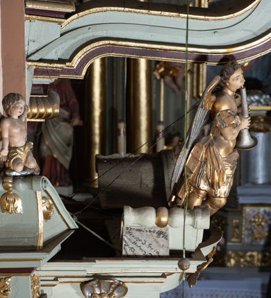 Zdjęcie nr 13: Ambona przyścienna, wisząca, z dostępem od zakrystii. Kosz na rzucie prostokąta z wypukłą ścianką frontową, z dodatkowym podestem od strony prezbiterium, ustawiony na niewielkiej podstawie dekorowanej muszlą, z podwieszeniem w formie zwężającego się cokołu ozdobionego ornamentem roślinnym, zakończonym szyszką. Ścianki kosza dekorowane płycinami w kształcie kwadratu o wklęsłych narożach wypełnionymi dekoracją złożoną z akantu, rozet i kratki regencyjnej. Naroża ścianki przedniej zdobione zwisami utworzonymi z wstęgi i kratki regencyjnej oraz ornamentu roślinnego. Parapet w formie gzymsu. Zaplecek w kształcie pionowego prostokąta, dekorowany w polu profilowaną płyciną z ornamentem roślinnym i płaskorzeźbioną figurą św. Stanisława. Baldachim powtarzający plan kosza, ujęty niepełnym belkowaniem, dekorowanym z trzech stron szczytami, z fragmentami zwróconego na zewnątrz przerwanego, wolutowego przyczółka, stanowiący podstawę dla figury anioła. Na podniebieniu baldachimu gołębica Ducha Świętego w glorii promienistej. Struktura polichromowana w kolorze jasnozielonym ze złoconymi i srebrzonymi profilami oraz ornamentyką. 
Figura anioła pełnoplastyczna, zwrócona jest w trzech czwartych w prawo, w silnym kontrapoście, o esowato wygiętej sylwetce, z głową skierowaną w górę. Obiema rękami obejmuje trąbę, na której gra. Twarz ma pociągłą, o dużych oczach skierowanych w górę, zadartym nosie i pełnych ustach, okoloną silnie kręconymi włosami. Anioł ubrany jest w zapinaną z przodu na guziki, obcisłą kamizelkę z bufiastymi, krótkimi rękawami i wysokim kołnierzem, w pludry i pończochy, wokół bioder ma zawieszony płaszcz, podwiązany po bokach. Skrzydła duże, złożone po bokach postaci. Polichromia ciała naturalistyczna, strój złocony, ze srebrzonymi detalami. 
Po wewnętrznej stronie fragmentu lewego przerwanego przyczółka napis: „odnowiono 1841. / ołtarze 3. tęcę i ambonę / przez Malarza z Tar/nawy Andrzeja / Wierzch[…]”.