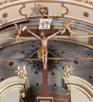 Zdjęcie nr 1: Grupa Ukrzyżowania ustawiona jest na belce tęczowej o falistym zarysie gzymsu z wyodrębnionym pośrodku cokołem z podwieszonym wazonem. Umieszczony na cokole krzyż jest flankowany fragmentami przerwanego półkolistego przyczółka z podwieszonymi wazonami. Do krzyża przybita jest trzema gwoździami pełnoplastyczna figura umarłego Chrystusa. Jego ciało przedstawiono w dużym zwisie, o układzie ramion w kształcie litery „V”, z głową opadającą na prawe ramię oraz stopami skrzyżowanymi w układzie prawa na lewą. Twarz ma pociągłą, o zamkniętych oczach, dużym nosie i otwartych ustach, okoloną krótką brodą oraz kręconymi w grube pukle włosami. Na głowie ma koronę cierniową. Ciało szczupłe o schematycznie zaznaczonych elementach anatomicznych. Perizonium złocone, krótkie, zawiązane na prawym boku, ze zwisem tkaniny. Pionowa belka krzyża zakończona tabliczką z napisem „J.N. / R.J.”, podtrzymywaną przez nadlatujące z góry putto. Na ramionach poprzecznej belki krzyża dwa putta. Na wewnętrznych krawędziach przerwanego przyczółka flankującego krzyż, siedzą dwa putta, a na zewnętrznych ustawione są rzeźby Marii i św. Jana. Po lewej stronie krzyża pełnoplastyczna figura Marii zwróconej w trzech czwartych w lewo z uniesioną głową i splecionymi dłońmi. Ubrana jest w różową suknię ze złoconymi lamówkami i niebieski płaszcz z takimiż lamówkami zarzucony na głowę i ramiona, przewiązany z przodu postaci. Po prawej stronie krzyża pełnoplastyczna figura św. Jana zwróconego w trzech czwartych w prawo, z uniesioną głową i prawą ręką w geście błogosławieństwa, lewą zaś wyciągniętą w bok. Ma pełną twarz okoloną silnie kręconymi włosami sięgającymi ramion. Ubrany jest w zieloną tunikę ze złoconymi lamówkami oraz czerwony płaszcz z podobnymi lamówkami przewiązany diagonalnie przez lewe ramię. Polichromia ciała figur naturalistyczna. 

