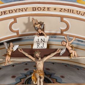 Zdjęcie nr 1: Grupa Ukrzyżowania ustawiona jest na belce tęczowej o falistym zarysie gzymsu z wyodrębnionym pośrodku cokołem z podwieszonym wazonem. Umieszczony na cokole krzyż jest flankowany fragmentami przerwanego półkolistego przyczółka z podwieszonymi wazonami. Do krzyża przybita jest trzema gwoździami pełnoplastyczna figura umarłego Chrystusa. Jego ciało przedstawiono w dużym zwisie, o układzie ramion w kształcie litery „V”, z głową opadającą na prawe ramię oraz stopami skrzyżowanymi w układzie prawa na lewą. Twarz ma pociągłą, o zamkniętych oczach, dużym nosie i otwartych ustach, okoloną krótką brodą oraz kręconymi w grube pukle włosami. Na głowie ma koronę cierniową. Ciało szczupłe o schematycznie zaznaczonych elementach anatomicznych. Perizonium złocone, krótkie, zawiązane na prawym boku, ze zwisem tkaniny. Pionowa belka krzyża zakończona tabliczką z napisem „J.N. / R.J.”, podtrzymywaną przez nadlatujące z góry putto. Na ramionach poprzecznej belki krzyża dwa putta. Na wewnętrznych krawędziach przerwanego przyczółka flankującego krzyż, siedzą dwa putta, a na zewnętrznych ustawione są rzeźby Marii i św. Jana. Po lewej stronie krzyża pełnoplastyczna figura Marii zwróconej w trzech czwartych w lewo z uniesioną głową i splecionymi dłońmi. Ubrana jest w różową suknię ze złoconymi lamówkami i niebieski płaszcz z takimiż lamówkami zarzucony na głowę i ramiona, przewiązany z przodu postaci. Po prawej stronie krzyża pełnoplastyczna figura św. Jana zwróconego w trzech czwartych w prawo, z uniesioną głową i prawą ręką w geście błogosławieństwa, lewą zaś wyciągniętą w bok. Ma pełną twarz okoloną silnie kręconymi włosami sięgającymi ramion. Ubrany jest w zieloną tunikę ze złoconymi lamówkami oraz czerwony płaszcz z podobnymi lamówkami przewiązany diagonalnie przez lewe ramię. Polichromia ciała figur naturalistyczna. 

