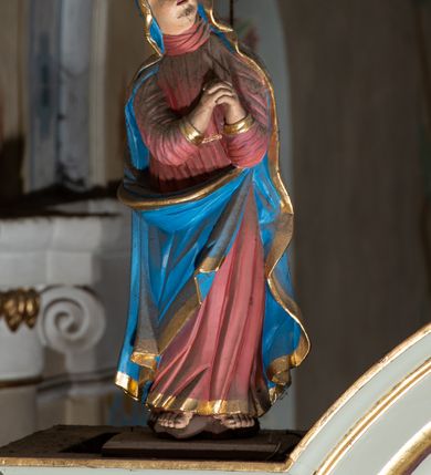 Zdjęcie nr 6: Grupa Ukrzyżowania ustawiona jest na belce tęczowej o falistym zarysie gzymsu z wyodrębnionym pośrodku cokołem z podwieszonym wazonem. Umieszczony na cokole krzyż jest flankowany fragmentami przerwanego półkolistego przyczółka z podwieszonymi wazonami. Do krzyża przybita jest trzema gwoździami pełnoplastyczna figura umarłego Chrystusa. Jego ciało przedstawiono w dużym zwisie, o układzie ramion w kształcie litery „V”, z głową opadającą na prawe ramię oraz stopami skrzyżowanymi w układzie prawa na lewą. Twarz ma pociągłą, o zamkniętych oczach, dużym nosie i otwartych ustach, okoloną krótką brodą oraz kręconymi w grube pukle włosami. Na głowie ma koronę cierniową. Ciało szczupłe o schematycznie zaznaczonych elementach anatomicznych. Perizonium złocone, krótkie, zawiązane na prawym boku, ze zwisem tkaniny. Pionowa belka krzyża zakończona tabliczką z napisem „J.N. / R.J.”, podtrzymywaną przez nadlatujące z góry putto. Na ramionach poprzecznej belki krzyża dwa putta. Na wewnętrznych krawędziach przerwanego przyczółka flankującego krzyż, siedzą dwa putta, a na zewnętrznych ustawione są rzeźby Marii i św. Jana. Po lewej stronie krzyża pełnoplastyczna figura Marii zwróconej w trzech czwartych w lewo z uniesioną głową i splecionymi dłońmi. Ubrana jest w różową suknię ze złoconymi lamówkami i niebieski płaszcz z takimiż lamówkami zarzucony na głowę i ramiona, przewiązany z przodu postaci. Po prawej stronie krzyża pełnoplastyczna figura św. Jana zwróconego w trzech czwartych w prawo, z uniesioną głową i prawą ręką w geście błogosławieństwa, lewą zaś wyciągniętą w bok. Ma pełną twarz okoloną silnie kręconymi włosami sięgającymi ramion. Ubrany jest w zieloną tunikę ze złoconymi lamówkami oraz czerwony płaszcz z podobnymi lamówkami przewiązany diagonalnie przez lewe ramię. Polichromia ciała figur naturalistyczna. 

