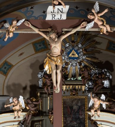 Zdjęcie nr 3: Grupa Ukrzyżowania ustawiona jest na belce tęczowej o falistym zarysie gzymsu z wyodrębnionym pośrodku cokołem z podwieszonym wazonem. Umieszczony na cokole krzyż jest flankowany fragmentami przerwanego półkolistego przyczółka z podwieszonymi wazonami. Do krzyża przybita jest trzema gwoździami pełnoplastyczna figura umarłego Chrystusa. Jego ciało przedstawiono w dużym zwisie, o układzie ramion w kształcie litery „V”, z głową opadającą na prawe ramię oraz stopami skrzyżowanymi w układzie prawa na lewą. Twarz ma pociągłą, o zamkniętych oczach, dużym nosie i otwartych ustach, okoloną krótką brodą oraz kręconymi w grube pukle włosami. Na głowie ma koronę cierniową. Ciało szczupłe o schematycznie zaznaczonych elementach anatomicznych. Perizonium złocone, krótkie, zawiązane na prawym boku, ze zwisem tkaniny. Pionowa belka krzyża zakończona tabliczką z napisem „J.N. / R.J.”, podtrzymywaną przez nadlatujące z góry putto. Na ramionach poprzecznej belki krzyża dwa putta. Na wewnętrznych krawędziach przerwanego przyczółka flankującego krzyż, siedzą dwa putta, a na zewnętrznych ustawione są rzeźby Marii i św. Jana. Po lewej stronie krzyża pełnoplastyczna figura Marii zwróconej w trzech czwartych w lewo z uniesioną głową i splecionymi dłońmi. Ubrana jest w różową suknię ze złoconymi lamówkami i niebieski płaszcz z takimiż lamówkami zarzucony na głowę i ramiona, przewiązany z przodu postaci. Po prawej stronie krzyża pełnoplastyczna figura św. Jana zwróconego w trzech czwartych w prawo, z uniesioną głową i prawą ręką w geście błogosławieństwa, lewą zaś wyciągniętą w bok. Ma pełną twarz okoloną silnie kręconymi włosami sięgającymi ramion. Ubrany jest w zieloną tunikę ze złoconymi lamówkami oraz czerwony płaszcz z podobnymi lamówkami przewiązany diagonalnie przez lewe ramię. Polichromia ciała figur naturalistyczna. 

