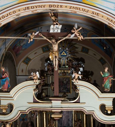Zdjęcie nr 2: Grupa Ukrzyżowania ustawiona jest na belce tęczowej o falistym zarysie gzymsu z wyodrębnionym pośrodku cokołem z podwieszonym wazonem. Umieszczony na cokole krzyż jest flankowany fragmentami przerwanego półkolistego przyczółka z podwieszonymi wazonami. Do krzyża przybita jest trzema gwoździami pełnoplastyczna figura umarłego Chrystusa. Jego ciało przedstawiono w dużym zwisie, o układzie ramion w kształcie litery „V”, z głową opadającą na prawe ramię oraz stopami skrzyżowanymi w układzie prawa na lewą. Twarz ma pociągłą, o zamkniętych oczach, dużym nosie i otwartych ustach, okoloną krótką brodą oraz kręconymi w grube pukle włosami. Na głowie ma koronę cierniową. Ciało szczupłe o schematycznie zaznaczonych elementach anatomicznych. Perizonium złocone, krótkie, zawiązane na prawym boku, ze zwisem tkaniny. Pionowa belka krzyża zakończona tabliczką z napisem „J.N. / R.J.”, podtrzymywaną przez nadlatujące z góry putto. Na ramionach poprzecznej belki krzyża dwa putta. Na wewnętrznych krawędziach przerwanego przyczółka flankującego krzyż, siedzą dwa putta, a na zewnętrznych ustawione są rzeźby Marii i św. Jana. Po lewej stronie krzyża pełnoplastyczna figura Marii zwróconej w trzech czwartych w lewo z uniesioną głową i splecionymi dłońmi. Ubrana jest w różową suknię ze złoconymi lamówkami i niebieski płaszcz z takimiż lamówkami zarzucony na głowę i ramiona, przewiązany z przodu postaci. Po prawej stronie krzyża pełnoplastyczna figura św. Jana zwróconego w trzech czwartych w prawo, z uniesioną głową i prawą ręką w geście błogosławieństwa, lewą zaś wyciągniętą w bok. Ma pełną twarz okoloną silnie kręconymi włosami sięgającymi ramion. Ubrany jest w zieloną tunikę ze złoconymi lamówkami oraz czerwony płaszcz z podobnymi lamówkami przewiązany diagonalnie przez lewe ramię. Polichromia ciała figur naturalistyczna. 


