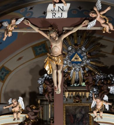 Zdjęcie nr 4: Grupa Ukrzyżowania ustawiona jest na belce tęczowej o falistym zarysie gzymsu z wyodrębnionym pośrodku cokołem z podwieszonym wazonem. Umieszczony na cokole krzyż jest flankowany fragmentami przerwanego półkolistego przyczółka z podwieszonymi wazonami. Do krzyża przybita jest trzema gwoździami pełnoplastyczna figura umarłego Chrystusa. Jego ciało przedstawiono w dużym zwisie, o układzie ramion w kształcie litery „V”, z głową opadającą na prawe ramię oraz stopami skrzyżowanymi w układzie prawa na lewą. Twarz ma pociągłą, o zamkniętych oczach, dużym nosie i otwartych ustach, okoloną krótką brodą oraz kręconymi w grube pukle włosami. Na głowie ma koronę cierniową. Ciało szczupłe o schematycznie zaznaczonych elementach anatomicznych. Perizonium złocone, krótkie, zawiązane na prawym boku, ze zwisem tkaniny. Pionowa belka krzyża zakończona tabliczką z napisem „J.N. / R.J.”, podtrzymywaną przez nadlatujące z góry putto. Na ramionach poprzecznej belki krzyża dwa putta. Na wewnętrznych krawędziach przerwanego przyczółka flankującego krzyż, siedzą dwa putta, a na zewnętrznych ustawione są rzeźby Marii i św. Jana. Po lewej stronie krzyża pełnoplastyczna figura Marii zwróconej w trzech czwartych w lewo z uniesioną głową i splecionymi dłońmi. Ubrana jest w różową suknię ze złoconymi lamówkami i niebieski płaszcz z takimiż lamówkami zarzucony na głowę i ramiona, przewiązany z przodu postaci. Po prawej stronie krzyża pełnoplastyczna figura św. Jana zwróconego w trzech czwartych w prawo, z uniesioną głową i prawą ręką w geście błogosławieństwa, lewą zaś wyciągniętą w bok. Ma pełną twarz okoloną silnie kręconymi włosami sięgającymi ramion. Ubrany jest w zieloną tunikę ze złoconymi lamówkami oraz czerwony płaszcz z podobnymi lamówkami przewiązany diagonalnie przez lewe ramię. Polichromia ciała figur naturalistyczna. 

