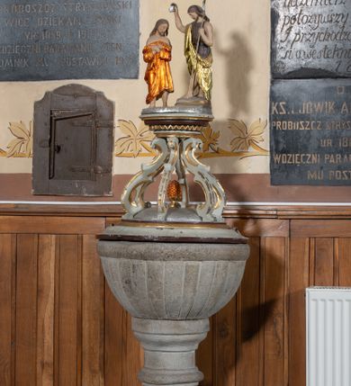 Zdjęcie nr 1: Chrzcielnica kamienna, ustawiona na dwustopniowej podstawie, na planie półkola. Stopa na planie koła, na wysokim cokole, gładka. Trzon prosty, nodus gruszkowaty, ujęty dwoma pierścieniami. Podstawa czary wklęsło-wypukła. Czara półkolista z imitacją puklowania, z wyodrębnionym brzegiem. Pokrywa drewniana, profilowana, z sześcioma kabłąkami podtrzymującymi cokół, na którym stoi grupa rzeźbiarska Chrztu Chrystusa. 


 