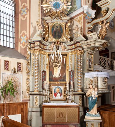 Zdjęcie nr 2: Nastawa architektoniczna, jednoosiowa, jednokondygnacyjna ze zwieńczeniem, ustawiona na cokole z wyodrębnionymi po bokach dwoma postumentami. Ołtarz prostopadłościenny z drewnianą mensą, z portatylem. Antepedium z kwiecistej tkaniny, z wyhaftowanym monogramem Marii w centrum, ujęte profilowaną ramą. Nastawa flankowana parą kolumn o skręconych trzonach i kapitelach kompozytowych stojących na tle dwóch par pilastrów o trzonach dekorowanych kampanulami. Podpory dźwigają pełne belkowanie, wyłamane na ich osi, o wklęsłym zarysie, przerwane nad polem środkowym, z fryzem dekorowanym rozetami oraz fragmenty przerwanego, falistego przyczółka ozdobionego kampanulami z parą puttów z wachlarzami. W polu nisza w formie stojącego prostokąta z obrazem Matki Boskiej Niepokalanie Poczętej w profilowanej ramie, flankowana dwiema gablotami z wotami. Nad polem środkowym dwa putta trzymające koronę zamkniętą oraz ryngraf malowany na blasze z przedstawieniem Matki Boskiej Bolesnej. Nastawa flankowana uszami utworzonymi z akantu i wstęgi regencyjnej. Zwieńczenie w formie stojącego prostokąta, o wklęsłych bokach, zamkniętego odcinkiem gzymsu wyłamanym w łuk ostry, ujęte liśćmi palmowymi z wstęgą. Całość zamknięta medalionem z monogramem Marii, w otoku z obłoków i uskrzydlonych główek anielskich, w glorii promienistej. W polu obraz przedstawiający Rodzinę Marii w formie stojącego owalu, w profilowanej ramie. Pole zwieńczenia wypełnione ornamentem roślinnym. Struktura architektoniczna polichromowana w kolorze jasnozielonym, kolumny, ornamentyka i profile złocone i srebrzone. 