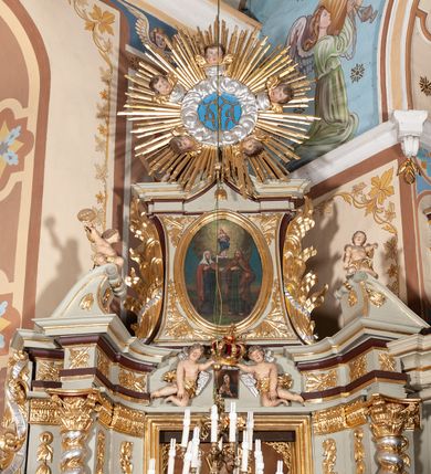 Zdjęcie nr 5: Nastawa architektoniczna, jednoosiowa, jednokondygnacyjna ze zwieńczeniem, ustawiona na cokole z wyodrębnionymi po bokach dwoma postumentami. Ołtarz prostopadłościenny z drewnianą mensą, z portatylem. Antepedium z kwiecistej tkaniny, z wyhaftowanym monogramem Marii w centrum, ujęte profilowaną ramą. Nastawa flankowana parą kolumn o skręconych trzonach i kapitelach kompozytowych stojących na tle dwóch par pilastrów o trzonach dekorowanych kampanulami. Podpory dźwigają pełne belkowanie, wyłamane na ich osi, o wklęsłym zarysie, przerwane nad polem środkowym, z fryzem dekorowanym rozetami oraz fragmenty przerwanego, falistego przyczółka ozdobionego kampanulami z parą puttów z wachlarzami. W polu nisza w formie stojącego prostokąta z obrazem Matki Boskiej Niepokalanie Poczętej w profilowanej ramie, flankowana dwiema gablotami z wotami. Nad polem środkowym dwa putta trzymające koronę zamkniętą oraz ryngraf malowany na blasze z przedstawieniem Matki Boskiej Bolesnej. Nastawa flankowana uszami utworzonymi z akantu i wstęgi regencyjnej. Zwieńczenie w formie stojącego prostokąta, o wklęsłych bokach, zamkniętego odcinkiem gzymsu wyłamanym w łuk ostry, ujęte liśćmi palmowymi z wstęgą. Całość zamknięta medalionem z monogramem Marii, w otoku z obłoków i uskrzydlonych główek anielskich, w glorii promienistej. W polu obraz przedstawiający Rodzinę Marii w formie stojącego owalu, w profilowanej ramie. Pole zwieńczenia wypełnione ornamentem roślinnym. Struktura architektoniczna polichromowana w kolorze jasnozielonym, kolumny, ornamentyka i profile złocone i srebrzone. 