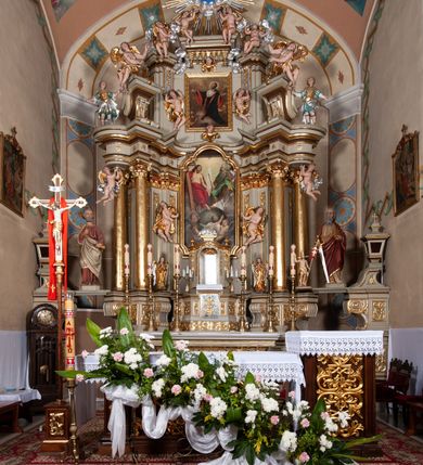 Zdjęcie nr 9: Nastawa architektoniczna, jednokondygnacyjna ze zwieńczeniem, jednoosiowa z bramkami. Ołtarz sarkofagowy z drewnianą mensą i portatylem. Na mensie znajduje się tabernakulum z tronem wystawienia. Tabernakulum w formie drewnianej skrzynki na planie wieloboku, ustawionej na cokole dekorowanym akantem, zamkniętej profilowaną listwą; ścianki dekorowane płycinami z akantem. Drzwiczki drewniane, obite blachą, w formie stojącego prostokąta, zdobione motywem krzyża, gorejącego serca, winnego grona i kłosów zboża. Tron wystawienia w formie obrotowej niszy, ujętej motywem winnego grona, flankowanej pilastrami dekorowanymi kampanulami, dźwigającymi gzyms zamykający całość, z wazonikami. Tron zwieńczony cokołem ze srebrzoną figurką leżącego baranka. 
Retabulum na dwustrefowym, uskokowym cokole ustawionym pod kątem, w górnej strefie z wyodrębnionymi po bokach postumentami i konsolami. Nastawa ujęta dwiema parami kolumn kompozytowych ustawionych na tle uskokowych, zwróconych pod kątem filarów, o trzonach dekorowanych płycinami z kampanulami. Podpory oraz dwa putta dźwigają wyłamane na ich osi, odcinkami wklęsło-wypukłe, pełne belkowanie, przerwane nad polem środkowym, z fryzem dekorowanym rozetami oraz fragmenty przerwanego, falistego przyczółka z siedzącymi na nim figurami aniołów. Na zewnętrznych krawędziach belkowania ustawione dwie figury w strojach żołnierzy. W polu środkowym nisza w formie stojącego prostokąta zamkniętego trójlistnie z uskokiem, o wklęsłych dolnych narożach, ujęta profilowaną ramą, z obrazem Trójcy Świętej i flankowana parą puttów. 
Zwieńczenie w formie stojącego prostokąta, flankowane parą zdwojonych pilastrów, zwróconych na zewnątrz, zamknięte odcinkiem gzymsu dekorowanym lambrekinem, ujęte obłokami z dwiema parami siedzących puttów. Powyżej medalion z okiem Opatrzności, ujętym obłokami i uskrzydlonymi główkami anielskimi, w glorii promienistej. W polu zwieńczenia obraz św. Jana Kantego w formie stojącego prostokąta, ujęty profilowaną ramą. 
Na wysokości cokołu nastawę flankują bramki utworzone przez postumenty przylegające do ścian prezbiterium zwieńczone fragmentami falistego przyczółka z latarniami. Cokoły przy ścianie z nastawą łączą listwy, tworząc przejścia, nad którymi stoją figury św. Piotra i św. Pawła. 
Struktura polichromowana w kolorze jasnozielonym, z brązowymi detalami, czerwonymi kapitelami; kolumny, ornamentyka i profile złocone, gloria i obłoki srebrzone. 

