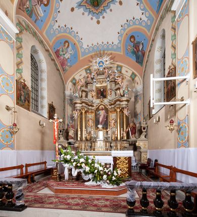 Zdjęcie nr 2: Nastawa architektoniczna, jednokondygnacyjna ze zwieńczeniem, jednoosiowa z bramkami. Ołtarz sarkofagowy z drewnianą mensą i portatylem. Na mensie znajduje się tabernakulum z tronem wystawienia. Tabernakulum w formie drewnianej skrzynki na planie wieloboku, ustawionej na cokole dekorowanym akantem, zamkniętej profilowaną listwą; ścianki dekorowane płycinami z akantem. Drzwiczki drewniane, obite blachą, w formie stojącego prostokąta, zdobione motywem krzyża, gorejącego serca, winnego grona i kłosów zboża. Tron wystawienia w formie obrotowej niszy, ujętej motywem winnego grona, flankowanej pilastrami dekorowanymi kampanulami, dźwigającymi gzyms zamykający całość, z wazonikami. Tron zwieńczony cokołem ze srebrzoną figurką leżącego baranka. 
Retabulum na dwustrefowym, uskokowym cokole ustawionym pod kątem, w górnej strefie z wyodrębnionymi po bokach postumentami i konsolami. Nastawa ujęta dwiema parami kolumn kompozytowych ustawionych na tle uskokowych, zwróconych pod kątem filarów, o trzonach dekorowanych płycinami z kampanulami. Podpory oraz dwa putta dźwigają wyłamane na ich osi, odcinkami wklęsło-wypukłe, pełne belkowanie, przerwane nad polem środkowym, z fryzem dekorowanym rozetami oraz fragmenty przerwanego, falistego przyczółka z siedzącymi na nim figurami aniołów. Na zewnętrznych krawędziach belkowania ustawione dwie figury w strojach żołnierzy. W polu środkowym nisza w formie stojącego prostokąta zamkniętego trójlistnie z uskokiem, o wklęsłych dolnych narożach, ujęta profilowaną ramą, z obrazem Trójcy Świętej i flankowana parą puttów. 
Zwieńczenie w formie stojącego prostokąta, flankowane parą zdwojonych pilastrów, zwróconych na zewnątrz, zamknięte odcinkiem gzymsu dekorowanym lambrekinem, ujęte obłokami z dwiema parami siedzących puttów. Powyżej medalion z okiem Opatrzności, ujętym obłokami i uskrzydlonymi główkami anielskimi, w glorii promienistej. W polu zwieńczenia obraz św. Jana Kantego w formie stojącego prostokąta, ujęty profilowaną ramą. 
Na wysokości cokołu nastawę flankują bramki utworzone przez postumenty przylegające do ścian prezbiterium zwieńczone fragmentami falistego przyczółka z latarniami. Cokoły przy ścianie z nastawą łączą listwy, tworząc przejścia, nad którymi stoją figury św. Piotra i św. Pawła. 
Struktura polichromowana w kolorze jasnozielonym, z brązowymi detalami, czerwonymi kapitelami; kolumny, ornamentyka i profile złocone, gloria i obłoki srebrzone. 

