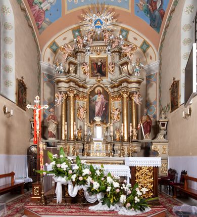 Zdjęcie nr 1: Nastawa architektoniczna, jednokondygnacyjna ze zwieńczeniem, jednoosiowa z bramkami. Ołtarz sarkofagowy z drewnianą mensą i portatylem. Na mensie znajduje się tabernakulum z tronem wystawienia. Tabernakulum w formie drewnianej skrzynki na planie wieloboku, ustawionej na cokole dekorowanym akantem, zamkniętej profilowaną listwą; ścianki dekorowane płycinami z akantem. Drzwiczki drewniane, obite blachą, w formie stojącego prostokąta, zdobione motywem krzyża, gorejącego serca, winnego grona i kłosów zboża. Tron wystawienia w formie obrotowej niszy, ujętej motywem winnego grona, flankowanej pilastrami dekorowanymi kampanulami, dźwigającymi gzyms zamykający całość, z wazonikami. Tron zwieńczony cokołem ze srebrzoną figurką leżącego baranka. 
Retabulum na dwustrefowym, uskokowym cokole ustawionym pod kątem, w górnej strefie z wyodrębnionymi po bokach postumentami i konsolami. Nastawa ujęta dwiema parami kolumn kompozytowych ustawionych na tle uskokowych, zwróconych pod kątem filarów, o trzonach dekorowanych płycinami z kampanulami. Podpory oraz dwa putta dźwigają wyłamane na ich osi, odcinkami wklęsło-wypukłe, pełne belkowanie, przerwane nad polem środkowym, z fryzem dekorowanym rozetami oraz fragmenty przerwanego, falistego przyczółka z siedzącymi na nim figurami aniołów. Na zewnętrznych krawędziach belkowania ustawione dwie figury w strojach żołnierzy. W polu środkowym nisza w formie stojącego prostokąta zamkniętego trójlistnie z uskokiem, o wklęsłych dolnych narożach, ujęta profilowaną ramą, z obrazem Trójcy Świętej i flankowana parą puttów. 
Zwieńczenie w formie stojącego prostokąta, flankowane parą zdwojonych pilastrów, zwróconych na zewnątrz, zamknięte odcinkiem gzymsu dekorowanym lambrekinem, ujęte obłokami z dwiema parami siedzących puttów. Powyżej medalion z okiem Opatrzności, ujętym obłokami i uskrzydlonymi główkami anielskimi, w glorii promienistej. W polu zwieńczenia obraz św. Jana Kantego w formie stojącego prostokąta, ujęty profilowaną ramą. 
Na wysokości cokołu nastawę flankują bramki utworzone przez postumenty przylegające do ścian prezbiterium zwieńczone fragmentami falistego przyczółka z latarniami. Cokoły przy ścianie z nastawą łączą listwy, tworząc przejścia, nad którymi stoją figury św. Piotra i św. Pawła. 
Struktura polichromowana w kolorze jasnozielonym, z brązowymi detalami, czerwonymi kapitelami; kolumny, ornamentyka i profile złocone, gloria i obłoki srebrzone. 

