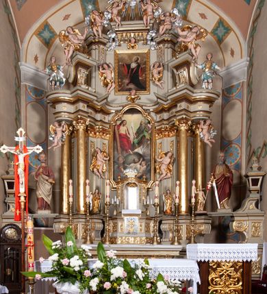 Zdjęcie nr 6: Nastawa architektoniczna, jednokondygnacyjna ze zwieńczeniem, jednoosiowa z bramkami. Ołtarz sarkofagowy z drewnianą mensą i portatylem. Na mensie znajduje się tabernakulum z tronem wystawienia. Tabernakulum w formie drewnianej skrzynki na planie wieloboku, ustawionej na cokole dekorowanym akantem, zamkniętej profilowaną listwą; ścianki dekorowane płycinami z akantem. Drzwiczki drewniane, obite blachą, w formie stojącego prostokąta, zdobione motywem krzyża, gorejącego serca, winnego grona i kłosów zboża. Tron wystawienia w formie obrotowej niszy, ujętej motywem winnego grona, flankowanej pilastrami dekorowanymi kampanulami, dźwigającymi gzyms zamykający całość, z wazonikami. Tron zwieńczony cokołem ze srebrzoną figurką leżącego baranka. 
Retabulum na dwustrefowym, uskokowym cokole ustawionym pod kątem, w górnej strefie z wyodrębnionymi po bokach postumentami i konsolami. Nastawa ujęta dwiema parami kolumn kompozytowych ustawionych na tle uskokowych, zwróconych pod kątem filarów, o trzonach dekorowanych płycinami z kampanulami. Podpory oraz dwa putta dźwigają wyłamane na ich osi, odcinkami wklęsło-wypukłe, pełne belkowanie, przerwane nad polem środkowym, z fryzem dekorowanym rozetami oraz fragmenty przerwanego, falistego przyczółka z siedzącymi na nim figurami aniołów. Na zewnętrznych krawędziach belkowania ustawione dwie figury w strojach żołnierzy. W polu środkowym nisza w formie stojącego prostokąta zamkniętego trójlistnie z uskokiem, o wklęsłych dolnych narożach, ujęta profilowaną ramą, z obrazem Trójcy Świętej i flankowana parą puttów. 
Zwieńczenie w formie stojącego prostokąta, flankowane parą zdwojonych pilastrów, zwróconych na zewnątrz, zamknięte odcinkiem gzymsu dekorowanym lambrekinem, ujęte obłokami z dwiema parami siedzących puttów. Powyżej medalion z okiem Opatrzności, ujętym obłokami i uskrzydlonymi główkami anielskimi, w glorii promienistej. W polu zwieńczenia obraz św. Jana Kantego w formie stojącego prostokąta, ujęty profilowaną ramą. 
Na wysokości cokołu nastawę flankują bramki utworzone przez postumenty przylegające do ścian prezbiterium zwieńczone fragmentami falistego przyczółka z latarniami. Cokoły przy ścianie z nastawą łączą listwy, tworząc przejścia, nad którymi stoją figury św. Piotra i św. Pawła. 
Struktura polichromowana w kolorze jasnozielonym, z brązowymi detalami, czerwonymi kapitelami; kolumny, ornamentyka i profile złocone, gloria i obłoki srebrzone. 

