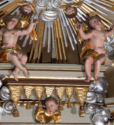 Zdjęcie nr 12: Nastawa architektoniczna, jednokondygnacyjna ze zwieńczeniem, jednoosiowa z bramkami. Ołtarz sarkofagowy z drewnianą mensą i portatylem. Na mensie znajduje się tabernakulum z tronem wystawienia. Tabernakulum w formie drewnianej skrzynki na planie wieloboku, ustawionej na cokole dekorowanym akantem, zamkniętej profilowaną listwą; ścianki dekorowane płycinami z akantem. Drzwiczki drewniane, obite blachą, w formie stojącego prostokąta, zdobione motywem krzyża, gorejącego serca, winnego grona i kłosów zboża. Tron wystawienia w formie obrotowej niszy, ujętej motywem winnego grona, flankowanej pilastrami dekorowanymi kampanulami, dźwigającymi gzyms zamykający całość, z wazonikami. Tron zwieńczony cokołem ze srebrzoną figurką leżącego baranka. 
Retabulum na dwustrefowym, uskokowym cokole ustawionym pod kątem, w górnej strefie z wyodrębnionymi po bokach postumentami i konsolami. Nastawa ujęta dwiema parami kolumn kompozytowych ustawionych na tle uskokowych, zwróconych pod kątem filarów, o trzonach dekorowanych płycinami z kampanulami. Podpory oraz dwa putta dźwigają wyłamane na ich osi, odcinkami wklęsło-wypukłe, pełne belkowanie, przerwane nad polem środkowym, z fryzem dekorowanym rozetami oraz fragmenty przerwanego, falistego przyczółka z siedzącymi na nim figurami aniołów. Na zewnętrznych krawędziach belkowania ustawione dwie figury w strojach żołnierzy. W polu środkowym nisza w formie stojącego prostokąta zamkniętego trójlistnie z uskokiem, o wklęsłych dolnych narożach, ujęta profilowaną ramą, z obrazem Trójcy Świętej i flankowana parą puttów. 
Zwieńczenie w formie stojącego prostokąta, flankowane parą zdwojonych pilastrów, zwróconych na zewnątrz, zamknięte odcinkiem gzymsu dekorowanym lambrekinem, ujęte obłokami z dwiema parami siedzących puttów. Powyżej medalion z okiem Opatrzności, ujętym obłokami i uskrzydlonymi główkami anielskimi, w glorii promienistej. W polu zwieńczenia obraz św. Jana Kantego w formie stojącego prostokąta, ujęty profilowaną ramą. 
Na wysokości cokołu nastawę flankują bramki utworzone przez postumenty przylegające do ścian prezbiterium zwieńczone fragmentami falistego przyczółka z latarniami. Cokoły przy ścianie z nastawą łączą listwy, tworząc przejścia, nad którymi stoją figury św. Piotra i św. Pawła. 
Struktura polichromowana w kolorze jasnozielonym, z brązowymi detalami, czerwonymi kapitelami; kolumny, ornamentyka i profile złocone, gloria i obłoki srebrzone. 

