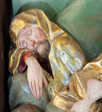 Zdjęcie nr 11: Płaskorzeźba o głębokim reliefie, w formie stojącego prostokąta z przedstawieniem modlącego się na skale Chrystusa w otoczeniu trzech śpiących apostołów. W centrum kompozycji postać klęczącego Chrystusa, zwróconego w trzech czwartych w lewo, z głową skierowaną niemal frontalnie oraz z dłońmi złożonymi w geście modlitwy. Twarz ma owalną, o wysokim czole, wąskich, oczach i nosie, małych ustach, okoloną krótką brodą i włosami rzeźbionymi w silnie skręcone pukle opadające na ramiona. Ubrany jest w luźną, laserowaną na czerwono tunikę, o szerokich, odwiniętych rękawach oraz złoty płaszcz zarzucony na lewy bok postaci, opadający z tyłu na nogi i z przodu na ziemię, silnie drapowany w łamane i misowe fałdy. Na pierwszym planie, w lewym dolnym rogu św. Jan ukazany jako śpiący, siedzący na lewej nodze, z ugiętą prawą przytrzymywaną obiema rękami przy korpusie, zwrócony jest w trzech czwartych w lewo, z głową opadającą na prawy bark. Twarz ma kwadratową, o zamkniętych, migdałowatych oczach, wąskim nosie i małych ustach, okoloną silnie kręconymi w rurkowate pukle włosami, sięgającymi ramion, z grzywką. Ubrany jest w czarną, luźną tunikę oraz czerwony płaszcz zarzucony na lewe ramię i zawinięty wokół nóg, przylegający do ciała. W prawym dolnym rogu śpiący św. Piotr, ukazany jako siedzący, z wysunięta do przodu lewą nogą, na prawej opiera łokieć ręki podtrzymującej jego opadającą głowę, lewa ręka spoczywa na kolanie. Zwrócony jest w trzech czwartych w prawo, z głową skierowana w przeciwną stronę. Twarz ma kwadratową, o zamkniętych, migdałowatych oczach, wąskim nosie i małych ustach, okoloną krótką, siwą silnie kręconą brodą oraz krótkimi włosami, z łysiną na głowie. Ubrany jest w granatową, luźną tunikę i srebrny płaszcz zarzucony na ramiona, zapięty pod szyją i opadający na nogi. Na trzecim planie, za skałą nad św. Janem wyłania się półpostać śpiącego św. Jakuba, opartego prawą ręką na skale, na której spoczywa jego głowa. Twarz ma kwadratową, o zamkniętych, migdałowatych oczach, wąskim nosie i małych ustach, okoloną krótką, brązową, silnie kręconą brodą. Ubrany jest w czerwoną, laserowaną tunikę i złocony płaszcz zarzucony na głowę i ramiona. W oddali, w lewym górnym rogu przy sztachetowym płocie widoczne są niewielkie trzy postacie Judasza i żołnierzy idących mostkiem. Tło w postaci zielonych pól. Płaskorzeźba ujęta profilowaną ramą. U spodu przedstawienia napis „Renovatum 1856 / Pinxyt Sitarski”.
