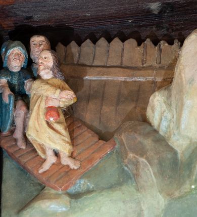 Zdjęcie nr 3: Płaskorzeźba o głębokim reliefie, w formie stojącego prostokąta z przedstawieniem modlącego się na skale Chrystusa w otoczeniu trzech śpiących apostołów. W centrum kompozycji postać klęczącego Chrystusa, zwróconego w trzech czwartych w lewo, z głową skierowaną niemal frontalnie oraz z dłońmi złożonymi w geście modlitwy. Twarz ma owalną, o wysokim czole, wąskich, oczach i nosie, małych ustach, okoloną krótką brodą i włosami rzeźbionymi w silnie skręcone pukle opadające na ramiona. Ubrany jest w luźną, laserowaną na czerwono tunikę, o szerokich, odwiniętych rękawach oraz złoty płaszcz zarzucony na lewy bok postaci, opadający z tyłu na nogi i z przodu na ziemię, silnie drapowany w łamane i misowe fałdy. Na pierwszym planie, w lewym dolnym rogu św. Jan ukazany jako śpiący, siedzący na lewej nodze, z ugiętą prawą przytrzymywaną obiema rękami przy korpusie, zwrócony jest w trzech czwartych w lewo, z głową opadającą na prawy bark. Twarz ma kwadratową, o zamkniętych, migdałowatych oczach, wąskim nosie i małych ustach, okoloną silnie kręconymi w rurkowate pukle włosami, sięgającymi ramion, z grzywką. Ubrany jest w czarną, luźną tunikę oraz czerwony płaszcz zarzucony na lewe ramię i zawinięty wokół nóg, przylegający do ciała. W prawym dolnym rogu śpiący św. Piotr, ukazany jako siedzący, z wysunięta do przodu lewą nogą, na prawej opiera łokieć ręki podtrzymującej jego opadającą głowę, lewa ręka spoczywa na kolanie. Zwrócony jest w trzech czwartych w prawo, z głową skierowana w przeciwną stronę. Twarz ma kwadratową, o zamkniętych, migdałowatych oczach, wąskim nosie i małych ustach, okoloną krótką, siwą silnie kręconą brodą oraz krótkimi włosami, z łysiną na głowie. Ubrany jest w granatową, luźną tunikę i srebrny płaszcz zarzucony na ramiona, zapięty pod szyją i opadający na nogi. Na trzecim planie, za skałą nad św. Janem wyłania się półpostać śpiącego św. Jakuba, opartego prawą ręką na skale, na której spoczywa jego głowa. Twarz ma kwadratową, o zamkniętych, migdałowatych oczach, wąskim nosie i małych ustach, okoloną krótką, brązową, silnie kręconą brodą. Ubrany jest w czerwoną, laserowaną tunikę i złocony płaszcz zarzucony na głowę i ramiona. W oddali, w lewym górnym rogu przy sztachetowym płocie widoczne są niewielkie trzy postacie Judasza i żołnierzy idących mostkiem. Tło w postaci zielonych pól. Płaskorzeźba ujęta profilowaną ramą. U spodu przedstawienia napis „Renovatum 1856 / Pinxyt Sitarski”.
