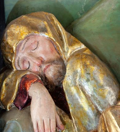 Zdjęcie nr 12: Płaskorzeźba o głębokim reliefie, w formie stojącego prostokąta z przedstawieniem modlącego się na skale Chrystusa w otoczeniu trzech śpiących apostołów. W centrum kompozycji postać klęczącego Chrystusa, zwróconego w trzech czwartych w lewo, z głową skierowaną niemal frontalnie oraz z dłońmi złożonymi w geście modlitwy. Twarz ma owalną, o wysokim czole, wąskich, oczach i nosie, małych ustach, okoloną krótką brodą i włosami rzeźbionymi w silnie skręcone pukle opadające na ramiona. Ubrany jest w luźną, laserowaną na czerwono tunikę, o szerokich, odwiniętych rękawach oraz złoty płaszcz zarzucony na lewy bok postaci, opadający z tyłu na nogi i z przodu na ziemię, silnie drapowany w łamane i misowe fałdy. Na pierwszym planie, w lewym dolnym rogu św. Jan ukazany jako śpiący, siedzący na lewej nodze, z ugiętą prawą przytrzymywaną obiema rękami przy korpusie, zwrócony jest w trzech czwartych w lewo, z głową opadającą na prawy bark. Twarz ma kwadratową, o zamkniętych, migdałowatych oczach, wąskim nosie i małych ustach, okoloną silnie kręconymi w rurkowate pukle włosami, sięgającymi ramion, z grzywką. Ubrany jest w czarną, luźną tunikę oraz czerwony płaszcz zarzucony na lewe ramię i zawinięty wokół nóg, przylegający do ciała. W prawym dolnym rogu śpiący św. Piotr, ukazany jako siedzący, z wysunięta do przodu lewą nogą, na prawej opiera łokieć ręki podtrzymującej jego opadającą głowę, lewa ręka spoczywa na kolanie. Zwrócony jest w trzech czwartych w prawo, z głową skierowana w przeciwną stronę. Twarz ma kwadratową, o zamkniętych, migdałowatych oczach, wąskim nosie i małych ustach, okoloną krótką, siwą silnie kręconą brodą oraz krótkimi włosami, z łysiną na głowie. Ubrany jest w granatową, luźną tunikę i srebrny płaszcz zarzucony na ramiona, zapięty pod szyją i opadający na nogi. Na trzecim planie, za skałą nad św. Janem wyłania się półpostać śpiącego św. Jakuba, opartego prawą ręką na skale, na której spoczywa jego głowa. Twarz ma kwadratową, o zamkniętych, migdałowatych oczach, wąskim nosie i małych ustach, okoloną krótką, brązową, silnie kręconą brodą. Ubrany jest w czerwoną, laserowaną tunikę i złocony płaszcz zarzucony na głowę i ramiona. W oddali, w lewym górnym rogu przy sztachetowym płocie widoczne są niewielkie trzy postacie Judasza i żołnierzy idących mostkiem. Tło w postaci zielonych pól. Płaskorzeźba ujęta profilowaną ramą. U spodu przedstawienia napis „Renovatum 1856 / Pinxyt Sitarski”.
