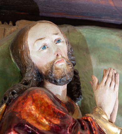 Zdjęcie nr 5: Płaskorzeźba o głębokim reliefie, w formie stojącego prostokąta z przedstawieniem modlącego się na skale Chrystusa w otoczeniu trzech śpiących apostołów. W centrum kompozycji postać klęczącego Chrystusa, zwróconego w trzech czwartych w lewo, z głową skierowaną niemal frontalnie oraz z dłońmi złożonymi w geście modlitwy. Twarz ma owalną, o wysokim czole, wąskich, oczach i nosie, małych ustach, okoloną krótką brodą i włosami rzeźbionymi w silnie skręcone pukle opadające na ramiona. Ubrany jest w luźną, laserowaną na czerwono tunikę, o szerokich, odwiniętych rękawach oraz złoty płaszcz zarzucony na lewy bok postaci, opadający z tyłu na nogi i z przodu na ziemię, silnie drapowany w łamane i misowe fałdy. Na pierwszym planie, w lewym dolnym rogu św. Jan ukazany jako śpiący, siedzący na lewej nodze, z ugiętą prawą przytrzymywaną obiema rękami przy korpusie, zwrócony jest w trzech czwartych w lewo, z głową opadającą na prawy bark. Twarz ma kwadratową, o zamkniętych, migdałowatych oczach, wąskim nosie i małych ustach, okoloną silnie kręconymi w rurkowate pukle włosami, sięgającymi ramion, z grzywką. Ubrany jest w czarną, luźną tunikę oraz czerwony płaszcz zarzucony na lewe ramię i zawinięty wokół nóg, przylegający do ciała. W prawym dolnym rogu śpiący św. Piotr, ukazany jako siedzący, z wysunięta do przodu lewą nogą, na prawej opiera łokieć ręki podtrzymującej jego opadającą głowę, lewa ręka spoczywa na kolanie. Zwrócony jest w trzech czwartych w prawo, z głową skierowana w przeciwną stronę. Twarz ma kwadratową, o zamkniętych, migdałowatych oczach, wąskim nosie i małych ustach, okoloną krótką, siwą silnie kręconą brodą oraz krótkimi włosami, z łysiną na głowie. Ubrany jest w granatową, luźną tunikę i srebrny płaszcz zarzucony na ramiona, zapięty pod szyją i opadający na nogi. Na trzecim planie, za skałą nad św. Janem wyłania się półpostać śpiącego św. Jakuba, opartego prawą ręką na skale, na której spoczywa jego głowa. Twarz ma kwadratową, o zamkniętych, migdałowatych oczach, wąskim nosie i małych ustach, okoloną krótką, brązową, silnie kręconą brodą. Ubrany jest w czerwoną, laserowaną tunikę i złocony płaszcz zarzucony na głowę i ramiona. W oddali, w lewym górnym rogu przy sztachetowym płocie widoczne są niewielkie trzy postacie Judasza i żołnierzy idących mostkiem. Tło w postaci zielonych pól. Płaskorzeźba ujęta profilowaną ramą. U spodu przedstawienia napis „Renovatum 1856 / Pinxyt Sitarski”.
