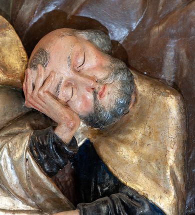 Zdjęcie nr 10: Płaskorzeźba o głębokim reliefie, w formie stojącego prostokąta z przedstawieniem modlącego się na skale Chrystusa w otoczeniu trzech śpiących apostołów. W centrum kompozycji postać klęczącego Chrystusa, zwróconego w trzech czwartych w lewo, z głową skierowaną niemal frontalnie oraz z dłońmi złożonymi w geście modlitwy. Twarz ma owalną, o wysokim czole, wąskich, oczach i nosie, małych ustach, okoloną krótką brodą i włosami rzeźbionymi w silnie skręcone pukle opadające na ramiona. Ubrany jest w luźną, laserowaną na czerwono tunikę, o szerokich, odwiniętych rękawach oraz złoty płaszcz zarzucony na lewy bok postaci, opadający z tyłu na nogi i z przodu na ziemię, silnie drapowany w łamane i misowe fałdy. Na pierwszym planie, w lewym dolnym rogu św. Jan ukazany jako śpiący, siedzący na lewej nodze, z ugiętą prawą przytrzymywaną obiema rękami przy korpusie, zwrócony jest w trzech czwartych w lewo, z głową opadającą na prawy bark. Twarz ma kwadratową, o zamkniętych, migdałowatych oczach, wąskim nosie i małych ustach, okoloną silnie kręconymi w rurkowate pukle włosami, sięgającymi ramion, z grzywką. Ubrany jest w czarną, luźną tunikę oraz czerwony płaszcz zarzucony na lewe ramię i zawinięty wokół nóg, przylegający do ciała. W prawym dolnym rogu śpiący św. Piotr, ukazany jako siedzący, z wysunięta do przodu lewą nogą, na prawej opiera łokieć ręki podtrzymującej jego opadającą głowę, lewa ręka spoczywa na kolanie. Zwrócony jest w trzech czwartych w prawo, z głową skierowana w przeciwną stronę. Twarz ma kwadratową, o zamkniętych, migdałowatych oczach, wąskim nosie i małych ustach, okoloną krótką, siwą silnie kręconą brodą oraz krótkimi włosami, z łysiną na głowie. Ubrany jest w granatową, luźną tunikę i srebrny płaszcz zarzucony na ramiona, zapięty pod szyją i opadający na nogi. Na trzecim planie, za skałą nad św. Janem wyłania się półpostać śpiącego św. Jakuba, opartego prawą ręką na skale, na której spoczywa jego głowa. Twarz ma kwadratową, o zamkniętych, migdałowatych oczach, wąskim nosie i małych ustach, okoloną krótką, brązową, silnie kręconą brodą. Ubrany jest w czerwoną, laserowaną tunikę i złocony płaszcz zarzucony na głowę i ramiona. W oddali, w lewym górnym rogu przy sztachetowym płocie widoczne są niewielkie trzy postacie Judasza i żołnierzy idących mostkiem. Tło w postaci zielonych pól. Płaskorzeźba ujęta profilowaną ramą. U spodu przedstawienia napis „Renovatum 1856 / Pinxyt Sitarski”.
