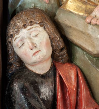 Zdjęcie nr 8: Płaskorzeźba o głębokim reliefie, w formie stojącego prostokąta z przedstawieniem modlącego się na skale Chrystusa w otoczeniu trzech śpiących apostołów. W centrum kompozycji postać klęczącego Chrystusa, zwróconego w trzech czwartych w lewo, z głową skierowaną niemal frontalnie oraz z dłońmi złożonymi w geście modlitwy. Twarz ma owalną, o wysokim czole, wąskich, oczach i nosie, małych ustach, okoloną krótką brodą i włosami rzeźbionymi w silnie skręcone pukle opadające na ramiona. Ubrany jest w luźną, laserowaną na czerwono tunikę, o szerokich, odwiniętych rękawach oraz złoty płaszcz zarzucony na lewy bok postaci, opadający z tyłu na nogi i z przodu na ziemię, silnie drapowany w łamane i misowe fałdy. Na pierwszym planie, w lewym dolnym rogu św. Jan ukazany jako śpiący, siedzący na lewej nodze, z ugiętą prawą przytrzymywaną obiema rękami przy korpusie, zwrócony jest w trzech czwartych w lewo, z głową opadającą na prawy bark. Twarz ma kwadratową, o zamkniętych, migdałowatych oczach, wąskim nosie i małych ustach, okoloną silnie kręconymi w rurkowate pukle włosami, sięgającymi ramion, z grzywką. Ubrany jest w czarną, luźną tunikę oraz czerwony płaszcz zarzucony na lewe ramię i zawinięty wokół nóg, przylegający do ciała. W prawym dolnym rogu śpiący św. Piotr, ukazany jako siedzący, z wysunięta do przodu lewą nogą, na prawej opiera łokieć ręki podtrzymującej jego opadającą głowę, lewa ręka spoczywa na kolanie. Zwrócony jest w trzech czwartych w prawo, z głową skierowana w przeciwną stronę. Twarz ma kwadratową, o zamkniętych, migdałowatych oczach, wąskim nosie i małych ustach, okoloną krótką, siwą silnie kręconą brodą oraz krótkimi włosami, z łysiną na głowie. Ubrany jest w granatową, luźną tunikę i srebrny płaszcz zarzucony na ramiona, zapięty pod szyją i opadający na nogi. Na trzecim planie, za skałą nad św. Janem wyłania się półpostać śpiącego św. Jakuba, opartego prawą ręką na skale, na której spoczywa jego głowa. Twarz ma kwadratową, o zamkniętych, migdałowatych oczach, wąskim nosie i małych ustach, okoloną krótką, brązową, silnie kręconą brodą. Ubrany jest w czerwoną, laserowaną tunikę i złocony płaszcz zarzucony na głowę i ramiona. W oddali, w lewym górnym rogu przy sztachetowym płocie widoczne są niewielkie trzy postacie Judasza i żołnierzy idących mostkiem. Tło w postaci zielonych pól. Płaskorzeźba ujęta profilowaną ramą. U spodu przedstawienia napis „Renovatum 1856 / Pinxyt Sitarski”.
