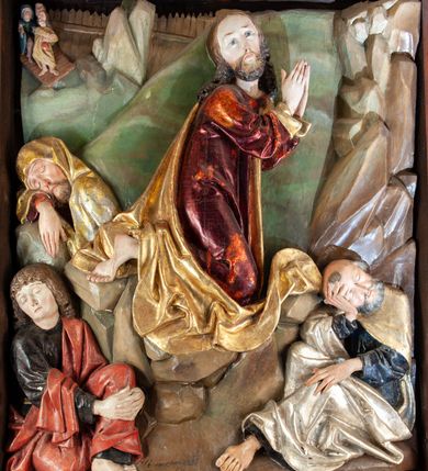 Zdjęcie nr 2: Płaskorzeźba o głębokim reliefie, w formie stojącego prostokąta z przedstawieniem modlącego się na skale Chrystusa w otoczeniu trzech śpiących apostołów. W centrum kompozycji postać klęczącego Chrystusa, zwróconego w trzech czwartych w lewo, z głową skierowaną niemal frontalnie oraz z dłońmi złożonymi w geście modlitwy. Twarz ma owalną, o wysokim czole, wąskich, oczach i nosie, małych ustach, okoloną krótką brodą i włosami rzeźbionymi w silnie skręcone pukle opadające na ramiona. Ubrany jest w luźną, laserowaną na czerwono tunikę, o szerokich, odwiniętych rękawach oraz złoty płaszcz zarzucony na lewy bok postaci, opadający z tyłu na nogi i z przodu na ziemię, silnie drapowany w łamane i misowe fałdy. Na pierwszym planie, w lewym dolnym rogu św. Jan ukazany jako śpiący, siedzący na lewej nodze, z ugiętą prawą przytrzymywaną obiema rękami przy korpusie, zwrócony jest w trzech czwartych w lewo, z głową opadającą na prawy bark. Twarz ma kwadratową, o zamkniętych, migdałowatych oczach, wąskim nosie i małych ustach, okoloną silnie kręconymi w rurkowate pukle włosami, sięgającymi ramion, z grzywką. Ubrany jest w czarną, luźną tunikę oraz czerwony płaszcz zarzucony na lewe ramię i zawinięty wokół nóg, przylegający do ciała. W prawym dolnym rogu śpiący św. Piotr, ukazany jako siedzący, z wysunięta do przodu lewą nogą, na prawej opiera łokieć ręki podtrzymującej jego opadającą głowę, lewa ręka spoczywa na kolanie. Zwrócony jest w trzech czwartych w prawo, z głową skierowana w przeciwną stronę. Twarz ma kwadratową, o zamkniętych, migdałowatych oczach, wąskim nosie i małych ustach, okoloną krótką, siwą silnie kręconą brodą oraz krótkimi włosami, z łysiną na głowie. Ubrany jest w granatową, luźną tunikę i srebrny płaszcz zarzucony na ramiona, zapięty pod szyją i opadający na nogi. Na trzecim planie, za skałą nad św. Janem wyłania się półpostać śpiącego św. Jakuba, opartego prawą ręką na skale, na której spoczywa jego głowa. Twarz ma kwadratową, o zamkniętych, migdałowatych oczach, wąskim nosie i małych ustach, okoloną krótką, brązową, silnie kręconą brodą. Ubrany jest w czerwoną, laserowaną tunikę i złocony płaszcz zarzucony na głowę i ramiona. W oddali, w lewym górnym rogu przy sztachetowym płocie widoczne są niewielkie trzy postacie Judasza i żołnierzy idących mostkiem. Tło w postaci zielonych pól. Płaskorzeźba ujęta profilowaną ramą. U spodu przedstawienia napis „Renovatum 1856 / Pinxyt Sitarski”.
