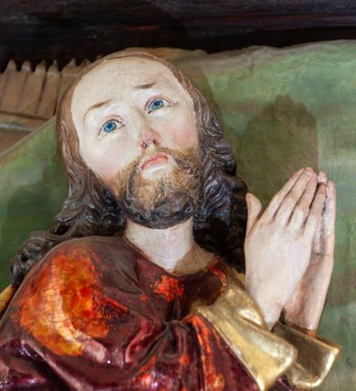 Zdjęcie nr 6: Płaskorzeźba o głębokim reliefie, w formie stojącego prostokąta z przedstawieniem modlącego się na skale Chrystusa w otoczeniu trzech śpiących apostołów. W centrum kompozycji postać klęczącego Chrystusa, zwróconego w trzech czwartych w lewo, z głową skierowaną niemal frontalnie oraz z dłońmi złożonymi w geście modlitwy. Twarz ma owalną, o wysokim czole, wąskich, oczach i nosie, małych ustach, okoloną krótką brodą i włosami rzeźbionymi w silnie skręcone pukle opadające na ramiona. Ubrany jest w luźną, laserowaną na czerwono tunikę, o szerokich, odwiniętych rękawach oraz złoty płaszcz zarzucony na lewy bok postaci, opadający z tyłu na nogi i z przodu na ziemię, silnie drapowany w łamane i misowe fałdy. Na pierwszym planie, w lewym dolnym rogu św. Jan ukazany jako śpiący, siedzący na lewej nodze, z ugiętą prawą przytrzymywaną obiema rękami przy korpusie, zwrócony jest w trzech czwartych w lewo, z głową opadającą na prawy bark. Twarz ma kwadratową, o zamkniętych, migdałowatych oczach, wąskim nosie i małych ustach, okoloną silnie kręconymi w rurkowate pukle włosami, sięgającymi ramion, z grzywką. Ubrany jest w czarną, luźną tunikę oraz czerwony płaszcz zarzucony na lewe ramię i zawinięty wokół nóg, przylegający do ciała. W prawym dolnym rogu śpiący św. Piotr, ukazany jako siedzący, z wysunięta do przodu lewą nogą, na prawej opiera łokieć ręki podtrzymującej jego opadającą głowę, lewa ręka spoczywa na kolanie. Zwrócony jest w trzech czwartych w prawo, z głową skierowana w przeciwną stronę. Twarz ma kwadratową, o zamkniętych, migdałowatych oczach, wąskim nosie i małych ustach, okoloną krótką, siwą silnie kręconą brodą oraz krótkimi włosami, z łysiną na głowie. Ubrany jest w granatową, luźną tunikę i srebrny płaszcz zarzucony na ramiona, zapięty pod szyją i opadający na nogi. Na trzecim planie, za skałą nad św. Janem wyłania się półpostać śpiącego św. Jakuba, opartego prawą ręką na skale, na której spoczywa jego głowa. Twarz ma kwadratową, o zamkniętych, migdałowatych oczach, wąskim nosie i małych ustach, okoloną krótką, brązową, silnie kręconą brodą. Ubrany jest w czerwoną, laserowaną tunikę i złocony płaszcz zarzucony na głowę i ramiona. W oddali, w lewym górnym rogu przy sztachetowym płocie widoczne są niewielkie trzy postacie Judasza i żołnierzy idących mostkiem. Tło w postaci zielonych pól. Płaskorzeźba ujęta profilowaną ramą. U spodu przedstawienia napis „Renovatum 1856 / Pinxyt Sitarski”.
