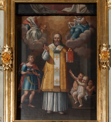 Zdjęcie nr 6: Obraz w formie stojącego prostokąta. W centrum przedstawienie św. Mikołaja z Jezusem i Marią unoszących się na obłokach w górnej części dzieła. Święty ukazany w całej postaci, frontalnie, lewą ręką podtrzymuje księgę z trzema kulami, prawą unosi z dłonią w geście błogosławieństwa. Twarz ma kwadratową z wysokim czołem, o starczych rysach, dużych oczach, prostym nosie i pełnych, rozchylonych ustach, okoloną siwą, kręcona brodą oraz siwymi, kręconymi włosami, sięgającymi ramion. Święty ubrany jest w strój biskupa wschodniego: biały sticharion, żółty sakkos z palicą przy boku oraz omoforion, na stopach ma buty. Po lewej stronie świętego znajduje się szaflik z trójką dzieci. Po jego prawej stronie postać młodzieńca, o pełnej, rumianej twarzy, okolonej jasnymi, kręconymi włosami, ubranego w niebieską tunikę sięgającą kolan oraz jasnoczerwony płaszcz zapięty na prawym ramieniu. Chłopiec trzyma tacę z karafką i dwoma kubkami. Postacie ukazane we wnętrzu. W górnej części obrazu centrycznie rozchodzące się, rozświetlone obłoki. W prawym górnym rogu siedząca na obłoku postać Chrystusa, skierowanego w trzech czwartych w lewo, który w lewej ręce trzyma księgę, a w prawej, opartej na globie, berło. Ubrany jest w czerwoną tunikę i biały płaszcz. Księgę podaje mu putto. W lewym górnym rogu siedząca na obłoku postać Marii, zwróconej w trzech czwartych w prawo, w obu wyciągniętych w przód dłoniach trzyma omoforion. Ubrana jest w jasnoróżową tunikę, przepasaną w talii, niebieski płaszcz i zielony welon. 
