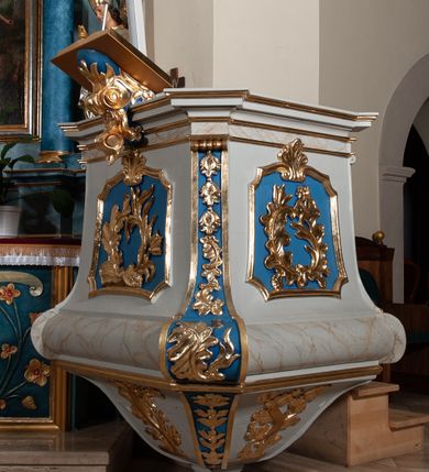 Zdjęcie nr 1: Ambona przyścienna, sięgająca ziemi, dostępna od strony prezbiterium. Kosz na planie kwadratu o ściętych narożach; na cokole w formie wałka. U spodu podwieszony sześcian zwężający się ku dołowi ze ściętymi narożami dekorowanymi płaskorzeźbionymi kampanulami; ścianki zdobione wstęgą w otoczeniu liści i kwiatów. Szyszka w kształcie profilowanego cokołu. Ścianki kosza wklęsłe, zwężające się ku górze; w narożach ujęte spływami wolutowymi; dekorowane płycinami o fantazyjnym zarysie zbliżonym do leżącego prostokąta z wklęsłymi narożami, zwieńczonymi wolutami z palmetą. W polach płaskorzeźbiona dekoracja roślinna w postaci liści, palm i kwiatów. Narożne woluty dekorowane kampanulami oraz liśćmi. Kosz zamknięty parapetem w formie niepełnego belkowania, wyłamanego w narożach. Od frontu pulpit oparty na wolucie i półkuli, dekorowanej liśćmi i kwiatami. Baldachim na planie kwadratu o ściętych narożach, którego podstawę stanowi niepełne belkowanie, wyłamane w narożach. Zwieńczenie utworzone z czterech kabłąków w formie wolut dekorowanych liściastymi festonami i wazonami; podtrzymujących cokół z rzeźbionym płomieniem. U spodu baldachimu w płycinie o formie kwadratu o wklęsłych narożach rzeźbiona gołębica Ducha Świętego w glorii promienistej. Struktura polichromowana w kolorze jasnoszarym, wałek i fryz z imitacją marmoryzacji z beżowymi żyłkami; płyciny i naroża niebieskie; ornamentyka, profilowania i kabłąki złocone.