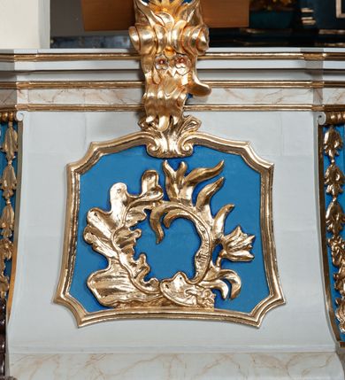 Zdjęcie nr 3: Ambona przyścienna, sięgająca ziemi, dostępna od strony prezbiterium. Kosz na planie kwadratu o ściętych narożach; na cokole w formie wałka. U spodu podwieszony sześcian zwężający się ku dołowi ze ściętymi narożami dekorowanymi płaskorzeźbionymi kampanulami; ścianki zdobione wstęgą w otoczeniu liści i kwiatów. Szyszka w kształcie profilowanego cokołu. Ścianki kosza wklęsłe, zwężające się ku górze; w narożach ujęte spływami wolutowymi; dekorowane płycinami o fantazyjnym zarysie zbliżonym do leżącego prostokąta z wklęsłymi narożami, zwieńczonymi wolutami z palmetą. W polach płaskorzeźbiona dekoracja roślinna w postaci liści, palm i kwiatów. Narożne woluty dekorowane kampanulami oraz liśćmi. Kosz zamknięty parapetem w formie niepełnego belkowania, wyłamanego w narożach. Od frontu pulpit oparty na wolucie i półkuli, dekorowanej liśćmi i kwiatami. Baldachim na planie kwadratu o ściętych narożach, którego podstawę stanowi niepełne belkowanie, wyłamane w narożach. Zwieńczenie utworzone z czterech kabłąków w formie wolut dekorowanych liściastymi festonami i wazonami; podtrzymujących cokół z rzeźbionym płomieniem. U spodu baldachimu w płycinie o formie kwadratu o wklęsłych narożach rzeźbiona gołębica Ducha Świętego w glorii promienistej. Struktura polichromowana w kolorze jasnoszarym, wałek i fryz z imitacją marmoryzacji z beżowymi żyłkami; płyciny i naroża niebieskie; ornamentyka, profilowania i kabłąki złocone.