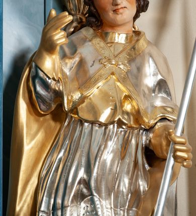 Zdjęcie nr 5: Rzeźba drążona, na cokole. Święty zwrócony frontalnie, w kontrapoście, o esowato wygiętej sylwetce, z głową przechyloną na prawe ramię. Prawą rękę unosi w geście błogosławieństwa, w lewej trzyma pastorał. Twarz ma pociągłą, oczy niewielkie, wydatny nos. Włosy długie, kręcone, opadające na ramiona. Jest ubrany w złoconą albę, srebrzoną komżę z koronką przy brzegu, złocony mucet z guzikami oraz srebrzony płaszcz zapięty na piersi, ze złoconą lamówką i podbiciem; na głowie ma złoconą infułę, na dłoniach rękawiczki. Polichromia naturalistyczna w odsłoniętych partiach ciała, strój i atrybuty złocone i srebrzone.