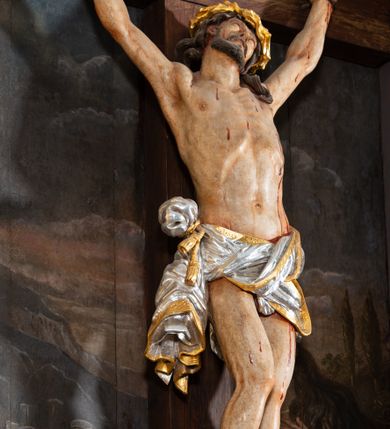 Zdjęcie nr 6: Pole główne ołtarza wypełnia figura Chrystusa Ukrzyżowanego na tle obrazu. Rzeźba pełna przedstawia żywego jeszcze Zbawiciela. Ciało oddano w dużym zwisie, o układzie ramion zbliżonym do litery „V” i esowato wygiętej sylwetce. Jezus ma nogi ugięte w kolanach, stopy przybite dwoma gwoździami, prawą nad lewą. Ciało szczupłe o wyraźnie podkreślonej muskulaturze i pracy mięśni, z zaznaczonymi śladami męki na lewym boku i w miejscach przebicia gwoździami. Głowa w grubej koronie cierniowej, przechylona na lewe ramię, uniesiona, ze wzrokiem skierowanym do góry. Twarz szczupła, okolona krótką brodą, nos długi, wąski, usta rozchylone. Włosy kręcone w pukle opadają na lewe ramię i plecy. Perizonium krótkie, utworzone ze skręconej, drapowanej i skrzyżowanej tkaniny, ze złoconą lamówką; przewiązane na prawym boku sznurem z chwostami, z obfitym, gęsto drapowanym zwisem sięgającym kolan. Polichromia naturalistyczna w partiach ciała; perizonium srebrzone i złocone, korona cierniowa i sznur złocone. W górnej części pionowej belki krzyża titulus w formie zwoju z napisem „IN/RI”. W tle, po prawej stronie krzyża postać klęczącej Marii Magdaleny, zwróconej w trzech czwartych w stronę Chrytsusa. Jej lewa ręka spoczywa na piersi, prawa ujmuje belkę krzyża. Ma pełną, młodzieńczą twarz, włosy długie, opadające na ramiona i plecy z wplecionymi sznurami pereł. Ubrana jest w białą suknię przewiązaną w talii oraz niebieski płaszcz połamany w grube fałdy na rękawach. U jej nóg puszka na wonności. W lewej dolnej części obrazu przedstawienie Jerozolimy wśród gór. Obraz w połowie przecina jaśniejsza linia horyzontu, powyżej widoczne ciemne niebo z kłębiastymi chmurami. Kolorystyka obrazu ciemna, kontrastująca z figurą Ukrzyżowanego. 