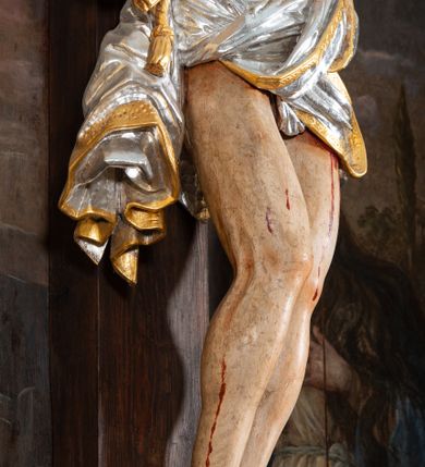 Zdjęcie nr 11: Pole główne ołtarza wypełnia figura Chrystusa Ukrzyżowanego na tle obrazu. Rzeźba pełna przedstawia żywego jeszcze Zbawiciela. Ciało oddano w dużym zwisie, o układzie ramion zbliżonym do litery „V” i esowato wygiętej sylwetce. Jezus ma nogi ugięte w kolanach, stopy przybite dwoma gwoździami, prawą nad lewą. Ciało szczupłe o wyraźnie podkreślonej muskulaturze i pracy mięśni, z zaznaczonymi śladami męki na lewym boku i w miejscach przebicia gwoździami. Głowa w grubej koronie cierniowej, przechylona na lewe ramię, uniesiona, ze wzrokiem skierowanym do góry. Twarz szczupła, okolona krótką brodą, nos długi, wąski, usta rozchylone. Włosy kręcone w pukle opadają na lewe ramię i plecy. Perizonium krótkie, utworzone ze skręconej, drapowanej i skrzyżowanej tkaniny, ze złoconą lamówką; przewiązane na prawym boku sznurem z chwostami, z obfitym, gęsto drapowanym zwisem sięgającym kolan. Polichromia naturalistyczna w partiach ciała; perizonium srebrzone i złocone, korona cierniowa i sznur złocone. W górnej części pionowej belki krzyża titulus w formie zwoju z napisem „IN/RI”. W tle, po prawej stronie krzyża postać klęczącej Marii Magdaleny, zwróconej w trzech czwartych w stronę Chrytsusa. Jej lewa ręka spoczywa na piersi, prawa ujmuje belkę krzyża. Ma pełną, młodzieńczą twarz, włosy długie, opadające na ramiona i plecy z wplecionymi sznurami pereł. Ubrana jest w białą suknię przewiązaną w talii oraz niebieski płaszcz połamany w grube fałdy na rękawach. U jej nóg puszka na wonności. W lewej dolnej części obrazu przedstawienie Jerozolimy wśród gór. Obraz w połowie przecina jaśniejsza linia horyzontu, powyżej widoczne ciemne niebo z kłębiastymi chmurami. Kolorystyka obrazu ciemna, kontrastująca z figurą Ukrzyżowanego. 