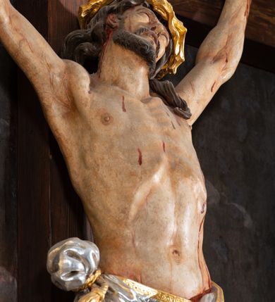 Zdjęcie nr 8: Pole główne ołtarza wypełnia figura Chrystusa Ukrzyżowanego na tle obrazu. Rzeźba pełna przedstawia żywego jeszcze Zbawiciela. Ciało oddano w dużym zwisie, o układzie ramion zbliżonym do litery „V” i esowato wygiętej sylwetce. Jezus ma nogi ugięte w kolanach, stopy przybite dwoma gwoździami, prawą nad lewą. Ciało szczupłe o wyraźnie podkreślonej muskulaturze i pracy mięśni, z zaznaczonymi śladami męki na lewym boku i w miejscach przebicia gwoździami. Głowa w grubej koronie cierniowej, przechylona na lewe ramię, uniesiona, ze wzrokiem skierowanym do góry. Twarz szczupła, okolona krótką brodą, nos długi, wąski, usta rozchylone. Włosy kręcone w pukle opadają na lewe ramię i plecy. Perizonium krótkie, utworzone ze skręconej, drapowanej i skrzyżowanej tkaniny, ze złoconą lamówką; przewiązane na prawym boku sznurem z chwostami, z obfitym, gęsto drapowanym zwisem sięgającym kolan. Polichromia naturalistyczna w partiach ciała; perizonium srebrzone i złocone, korona cierniowa i sznur złocone. W górnej części pionowej belki krzyża titulus w formie zwoju z napisem „IN/RI”. W tle, po prawej stronie krzyża postać klęczącej Marii Magdaleny, zwróconej w trzech czwartych w stronę Chrytsusa. Jej lewa ręka spoczywa na piersi, prawa ujmuje belkę krzyża. Ma pełną, młodzieńczą twarz, włosy długie, opadające na ramiona i plecy z wplecionymi sznurami pereł. Ubrana jest w białą suknię przewiązaną w talii oraz niebieski płaszcz połamany w grube fałdy na rękawach. U jej nóg puszka na wonności. W lewej dolnej części obrazu przedstawienie Jerozolimy wśród gór. Obraz w połowie przecina jaśniejsza linia horyzontu, powyżej widoczne ciemne niebo z kłębiastymi chmurami. Kolorystyka obrazu ciemna, kontrastująca z figurą Ukrzyżowanego. 