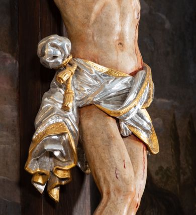 Zdjęcie nr 12: Pole główne ołtarza wypełnia figura Chrystusa Ukrzyżowanego na tle obrazu. Rzeźba pełna przedstawia żywego jeszcze Zbawiciela. Ciało oddano w dużym zwisie, o układzie ramion zbliżonym do litery „V” i esowato wygiętej sylwetce. Jezus ma nogi ugięte w kolanach, stopy przybite dwoma gwoździami, prawą nad lewą. Ciało szczupłe o wyraźnie podkreślonej muskulaturze i pracy mięśni, z zaznaczonymi śladami męki na lewym boku i w miejscach przebicia gwoździami. Głowa w grubej koronie cierniowej, przechylona na lewe ramię, uniesiona, ze wzrokiem skierowanym do góry. Twarz szczupła, okolona krótką brodą, nos długi, wąski, usta rozchylone. Włosy kręcone w pukle opadają na lewe ramię i plecy. Perizonium krótkie, utworzone ze skręconej, drapowanej i skrzyżowanej tkaniny, ze złoconą lamówką; przewiązane na prawym boku sznurem z chwostami, z obfitym, gęsto drapowanym zwisem sięgającym kolan. Polichromia naturalistyczna w partiach ciała; perizonium srebrzone i złocone, korona cierniowa i sznur złocone. W górnej części pionowej belki krzyża titulus w formie zwoju z napisem „IN/RI”. W tle, po prawej stronie krzyża postać klęczącej Marii Magdaleny, zwróconej w trzech czwartych w stronę Chrytsusa. Jej lewa ręka spoczywa na piersi, prawa ujmuje belkę krzyża. Ma pełną, młodzieńczą twarz, włosy długie, opadające na ramiona i plecy z wplecionymi sznurami pereł. Ubrana jest w białą suknię przewiązaną w talii oraz niebieski płaszcz połamany w grube fałdy na rękawach. U jej nóg puszka na wonności. W lewej dolnej części obrazu przedstawienie Jerozolimy wśród gór. Obraz w połowie przecina jaśniejsza linia horyzontu, powyżej widoczne ciemne niebo z kłębiastymi chmurami. Kolorystyka obrazu ciemna, kontrastująca z figurą Ukrzyżowanego. 