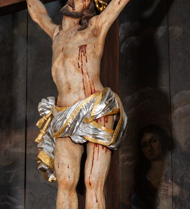 Zdjęcie nr 9: Pole główne ołtarza wypełnia figura Chrystusa Ukrzyżowanego na tle obrazu. Rzeźba pełna przedstawia żywego jeszcze Zbawiciela. Ciało oddano w dużym zwisie, o układzie ramion zbliżonym do litery „V” i esowato wygiętej sylwetce. Jezus ma nogi ugięte w kolanach, stopy przybite dwoma gwoździami, prawą nad lewą. Ciało szczupłe o wyraźnie podkreślonej muskulaturze i pracy mięśni, z zaznaczonymi śladami męki na lewym boku i w miejscach przebicia gwoździami. Głowa w grubej koronie cierniowej, przechylona na lewe ramię, uniesiona, ze wzrokiem skierowanym do góry. Twarz szczupła, okolona krótką brodą, nos długi, wąski, usta rozchylone. Włosy kręcone w pukle opadają na lewe ramię i plecy. Perizonium krótkie, utworzone ze skręconej, drapowanej i skrzyżowanej tkaniny, ze złoconą lamówką; przewiązane na prawym boku sznurem z chwostami, z obfitym, gęsto drapowanym zwisem sięgającym kolan. Polichromia naturalistyczna w partiach ciała; perizonium srebrzone i złocone, korona cierniowa i sznur złocone. W górnej części pionowej belki krzyża titulus w formie zwoju z napisem „IN/RI”. W tle, po prawej stronie krzyża postać klęczącej Marii Magdaleny, zwróconej w trzech czwartych w stronę Chrytsusa. Jej lewa ręka spoczywa na piersi, prawa ujmuje belkę krzyża. Ma pełną, młodzieńczą twarz, włosy długie, opadające na ramiona i plecy z wplecionymi sznurami pereł. Ubrana jest w białą suknię przewiązaną w talii oraz niebieski płaszcz połamany w grube fałdy na rękawach. U jej nóg puszka na wonności. W lewej dolnej części obrazu przedstawienie Jerozolimy wśród gór. Obraz w połowie przecina jaśniejsza linia horyzontu, powyżej widoczne ciemne niebo z kłębiastymi chmurami. Kolorystyka obrazu ciemna, kontrastująca z figurą Ukrzyżowanego. 