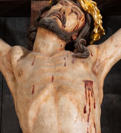 Zdjęcie nr 13: Pole główne ołtarza wypełnia figura Chrystusa Ukrzyżowanego na tle obrazu. Rzeźba pełna przedstawia żywego jeszcze Zbawiciela. Ciało oddano w dużym zwisie, o układzie ramion zbliżonym do litery „V” i esowato wygiętej sylwetce. Jezus ma nogi ugięte w kolanach, stopy przybite dwoma gwoździami, prawą nad lewą. Ciało szczupłe o wyraźnie podkreślonej muskulaturze i pracy mięśni, z zaznaczonymi śladami męki na lewym boku i w miejscach przebicia gwoździami. Głowa w grubej koronie cierniowej, przechylona na lewe ramię, uniesiona, ze wzrokiem skierowanym do góry. Twarz szczupła, okolona krótką brodą, nos długi, wąski, usta rozchylone. Włosy kręcone w pukle opadają na lewe ramię i plecy. Perizonium krótkie, utworzone ze skręconej, drapowanej i skrzyżowanej tkaniny, ze złoconą lamówką; przewiązane na prawym boku sznurem z chwostami, z obfitym, gęsto drapowanym zwisem sięgającym kolan. Polichromia naturalistyczna w partiach ciała; perizonium srebrzone i złocone, korona cierniowa i sznur złocone. W górnej części pionowej belki krzyża titulus w formie zwoju z napisem „IN/RI”. W tle, po prawej stronie krzyża postać klęczącej Marii Magdaleny, zwróconej w trzech czwartych w stronę Chrytsusa. Jej lewa ręka spoczywa na piersi, prawa ujmuje belkę krzyża. Ma pełną, młodzieńczą twarz, włosy długie, opadające na ramiona i plecy z wplecionymi sznurami pereł. Ubrana jest w białą suknię przewiązaną w talii oraz niebieski płaszcz połamany w grube fałdy na rękawach. U jej nóg puszka na wonności. W lewej dolnej części obrazu przedstawienie Jerozolimy wśród gór. Obraz w połowie przecina jaśniejsza linia horyzontu, powyżej widoczne ciemne niebo z kłębiastymi chmurami. Kolorystyka obrazu ciemna, kontrastująca z figurą Ukrzyżowanego. 