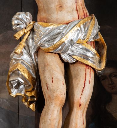 Zdjęcie nr 10: Pole główne ołtarza wypełnia figura Chrystusa Ukrzyżowanego na tle obrazu. Rzeźba pełna przedstawia żywego jeszcze Zbawiciela. Ciało oddano w dużym zwisie, o układzie ramion zbliżonym do litery „V” i esowato wygiętej sylwetce. Jezus ma nogi ugięte w kolanach, stopy przybite dwoma gwoździami, prawą nad lewą. Ciało szczupłe o wyraźnie podkreślonej muskulaturze i pracy mięśni, z zaznaczonymi śladami męki na lewym boku i w miejscach przebicia gwoździami. Głowa w grubej koronie cierniowej, przechylona na lewe ramię, uniesiona, ze wzrokiem skierowanym do góry. Twarz szczupła, okolona krótką brodą, nos długi, wąski, usta rozchylone. Włosy kręcone w pukle opadają na lewe ramię i plecy. Perizonium krótkie, utworzone ze skręconej, drapowanej i skrzyżowanej tkaniny, ze złoconą lamówką; przewiązane na prawym boku sznurem z chwostami, z obfitym, gęsto drapowanym zwisem sięgającym kolan. Polichromia naturalistyczna w partiach ciała; perizonium srebrzone i złocone, korona cierniowa i sznur złocone. W górnej części pionowej belki krzyża titulus w formie zwoju z napisem „IN/RI”. W tle, po prawej stronie krzyża postać klęczącej Marii Magdaleny, zwróconej w trzech czwartych w stronę Chrytsusa. Jej lewa ręka spoczywa na piersi, prawa ujmuje belkę krzyża. Ma pełną, młodzieńczą twarz, włosy długie, opadające na ramiona i plecy z wplecionymi sznurami pereł. Ubrana jest w białą suknię przewiązaną w talii oraz niebieski płaszcz połamany w grube fałdy na rękawach. U jej nóg puszka na wonności. W lewej dolnej części obrazu przedstawienie Jerozolimy wśród gór. Obraz w połowie przecina jaśniejsza linia horyzontu, powyżej widoczne ciemne niebo z kłębiastymi chmurami. Kolorystyka obrazu ciemna, kontrastująca z figurą Ukrzyżowanego. 