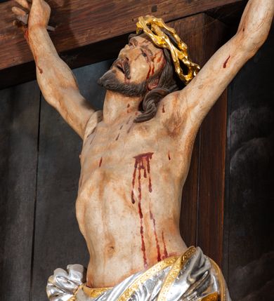 Zdjęcie nr 7: Pole główne ołtarza wypełnia figura Chrystusa Ukrzyżowanego na tle obrazu. Rzeźba pełna przedstawia żywego jeszcze Zbawiciela. Ciało oddano w dużym zwisie, o układzie ramion zbliżonym do litery „V” i esowato wygiętej sylwetce. Jezus ma nogi ugięte w kolanach, stopy przybite dwoma gwoździami, prawą nad lewą. Ciało szczupłe o wyraźnie podkreślonej muskulaturze i pracy mięśni, z zaznaczonymi śladami męki na lewym boku i w miejscach przebicia gwoździami. Głowa w grubej koronie cierniowej, przechylona na lewe ramię, uniesiona, ze wzrokiem skierowanym do góry. Twarz szczupła, okolona krótką brodą, nos długi, wąski, usta rozchylone. Włosy kręcone w pukle opadają na lewe ramię i plecy. Perizonium krótkie, utworzone ze skręconej, drapowanej i skrzyżowanej tkaniny, ze złoconą lamówką; przewiązane na prawym boku sznurem z chwostami, z obfitym, gęsto drapowanym zwisem sięgającym kolan. Polichromia naturalistyczna w partiach ciała; perizonium srebrzone i złocone, korona cierniowa i sznur złocone. W górnej części pionowej belki krzyża titulus w formie zwoju z napisem „IN/RI”. W tle, po prawej stronie krzyża postać klęczącej Marii Magdaleny, zwróconej w trzech czwartych w stronę Chrytsusa. Jej lewa ręka spoczywa na piersi, prawa ujmuje belkę krzyża. Ma pełną, młodzieńczą twarz, włosy długie, opadające na ramiona i plecy z wplecionymi sznurami pereł. Ubrana jest w białą suknię przewiązaną w talii oraz niebieski płaszcz połamany w grube fałdy na rękawach. U jej nóg puszka na wonności. W lewej dolnej części obrazu przedstawienie Jerozolimy wśród gór. Obraz w połowie przecina jaśniejsza linia horyzontu, powyżej widoczne ciemne niebo z kłębiastymi chmurami. Kolorystyka obrazu ciemna, kontrastująca z figurą Ukrzyżowanego. 