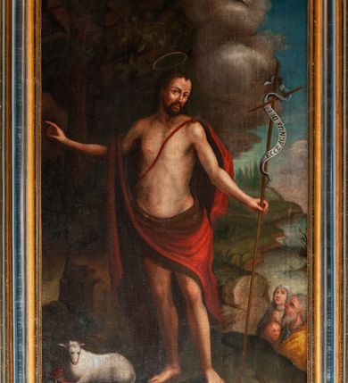 Zdjęcie nr 2: Obraz w kształcie stojącego prostokąta w profilowanej, złoconej i srebrzonej ramie z przedstawieniem św. Jana Chrzciciela. Święty stoi w centrum kompozycji w lekkim kontrapoście, skręcony delikatnie w prawo, z głową obróconą na lewe ramię. Prawą rękę unosi do boku, w lewej trzyma długie drzewce zakończone krzyżem z przewiązaną banderolą z napisem „ECCE AGNUS DEI”. Ma szczupłą, owalną twarz z wysokim czołem, wąskim nosem i dużymi, ciemnymi oczami. Nosi wąsy i krótką brodę, a długie włosy spływają mu na plecy. Święty ma narzucony czerwony, podbity brązem płaszcz przewiązany na lewym ramieniu szarfą, przerzucony przez prawe przedramię i otaczający biodra. U jego stóp, po lewej stronie kompozycji, na księdze w czerwonej oprawie leży niewielki baranek. Z prawej strony u dołu widać wznoszącą wzrok na świętego rodzinę złożoną ze starszego mężczyzny w niebieskiej sukni i żółtym płaszczu, kobiety w jasnych szatach i dziecka. Święty Jan został ukazany na tle ciemnego lasu lub bloku skalnego i skłębionego, szarego obłoku, ponad którym unosi się główka anielska. Przy prawej krawędzi widoczny fragment szerokiego krajobrazu stanowiącego brzeg rzeki lub morza. 