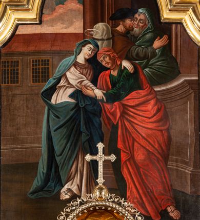 Zdjęcie nr 5: Obraz w formie stojącego prostokąta zamknięty łukiem półkolistym nadwieszonym z uskokami w złoconej, profilowanej ramie. W centrum postacie spotykających się Marii i św. Elżbiety. Kobiety zwrócone ku sobie obejmują się obiema rękami. Twarz Marii jest owalna o wyraźnych, młodzieńczych rysach, jasnej karnacji, nad głową nimb. Matka Boska jest ubrana w bladoróżową suknię przepasaną w talii oraz niebieski płaszcz nakrywający głowę i ramiona, przewiązany z przodu. Twarz św. Elżbiety jest owalna, zdradza jej starszy wiek, o ciemnej karnacji; oczy ma zwrócone w górę. Ubrana jest w zieloną suknię oraz czerwony płaszcz przerzucony przez prawe ramię i zawiązany na boku, na głowie ma jasnoczerwoną chustę. Na drugim planie w prawej części obrazu ukazany budynek, w którego wejściu przedstawione półpostacie witających się św. Józefa i Zachariasza. Scena została zakomponowana na tle architektury – na trzecim planie widoczny budynek z dwoma wielodzielnymi oknami. Powyżej w górnej części obrazu widoczny jest fragment błękitnego nieba z zabarwionymi na różowo obłokami. Kolorystyka ciemna, ciepła, postacie kobiet zostały wyróżnione intensywnym kolorem tkanin. Silny światłocień.